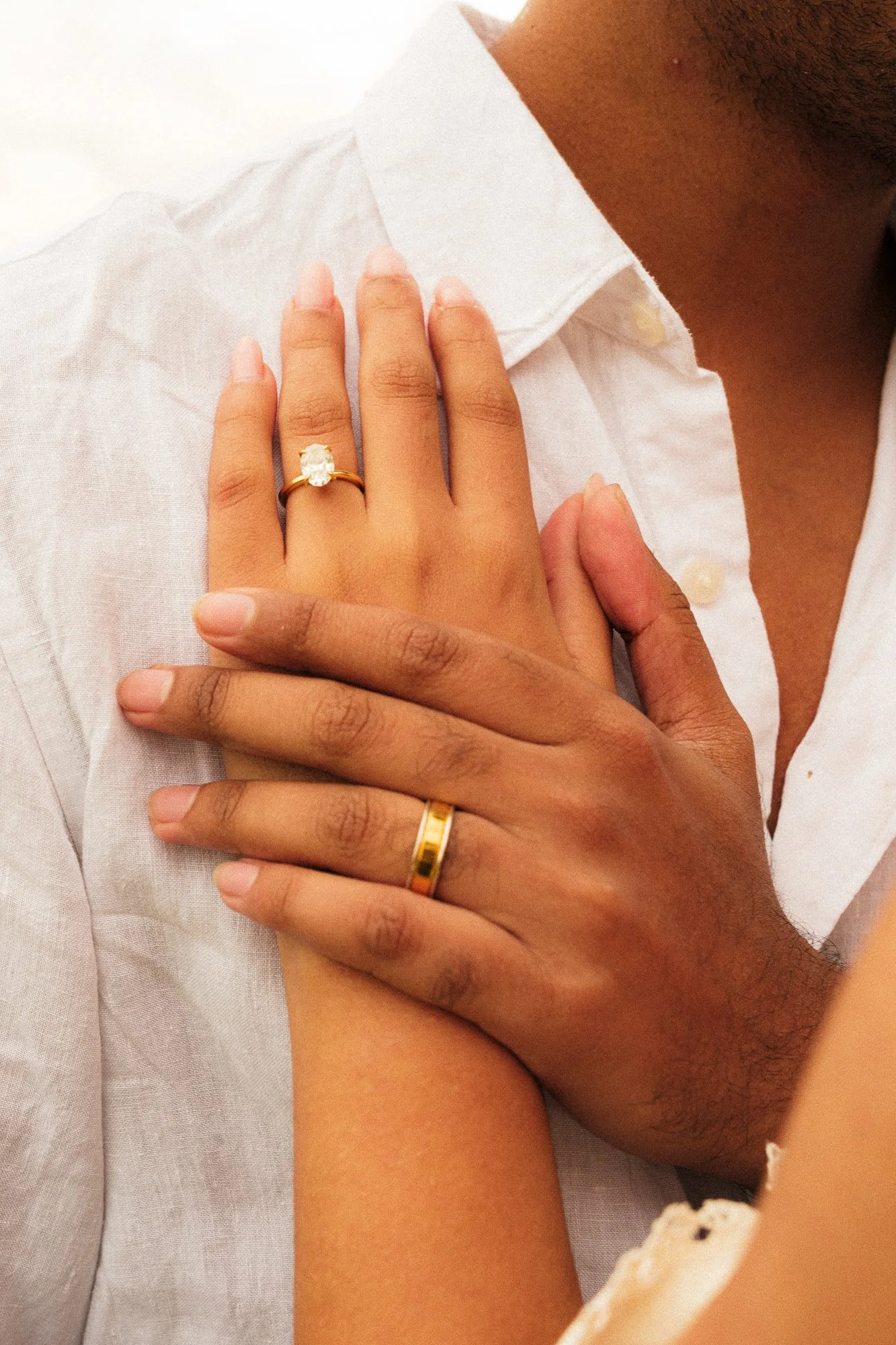 Close-up image of a couple's hands with wedding rings placed on each other's fingers, the woman wearing a gold ring with a large gemstone and the man wearing a gold band. The woman is wearing a white shirt, and the man's hand appears darker.