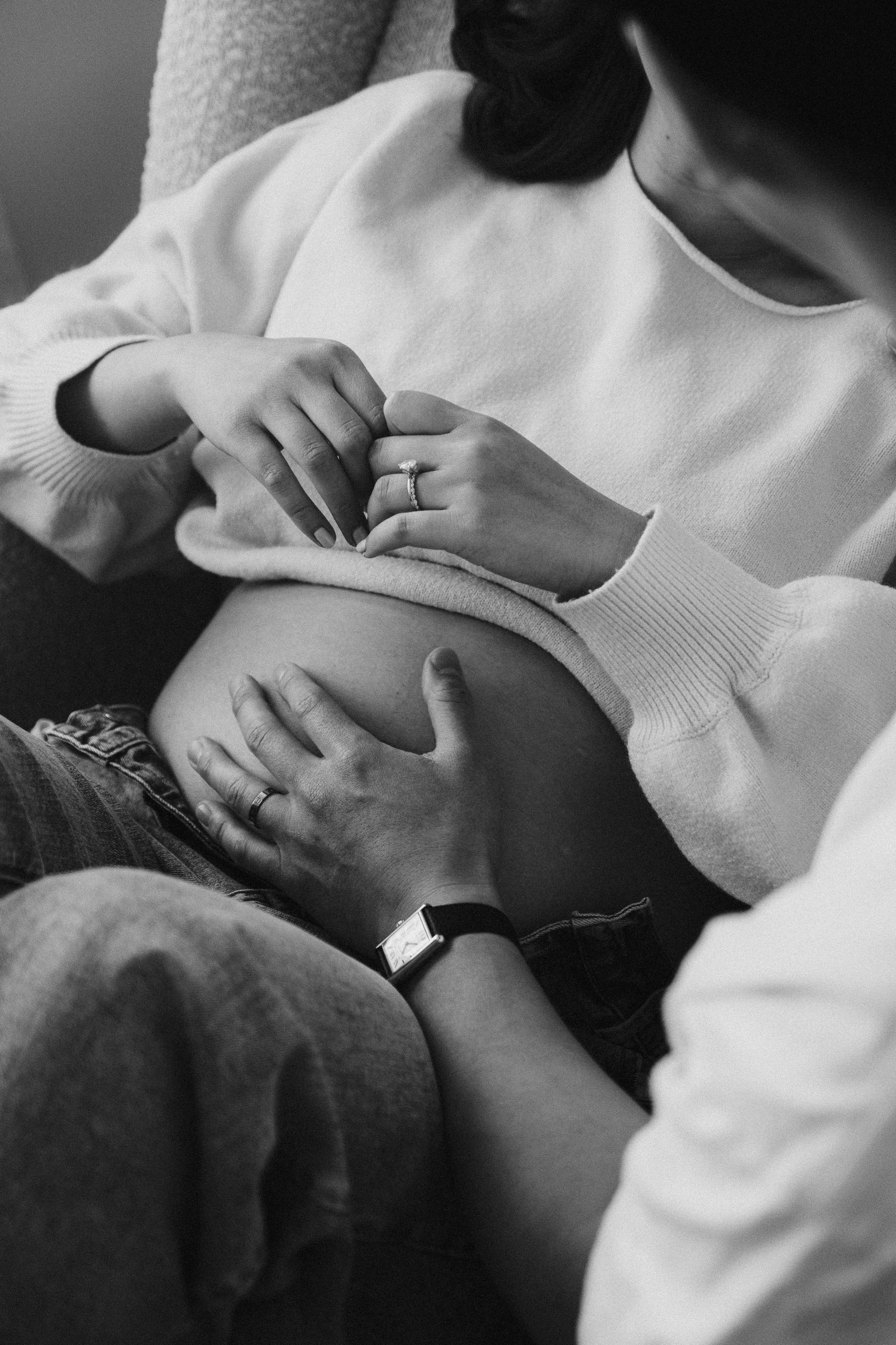 Black and white photo of a pregnant woman with a man, showing their hands on her belly. The woman is wearing a sweater and has a ring, the man is wearing a watch and a dark ring.