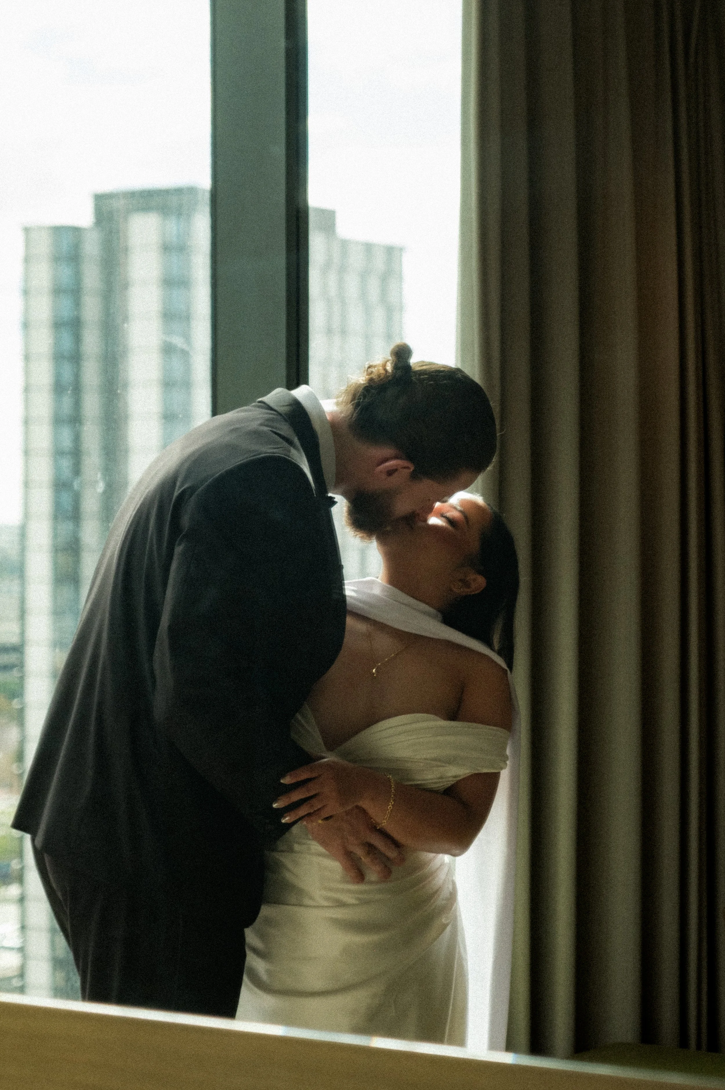 A man and woman sharing a intimate moment in front of a mirror in a room with large windows and cityscape view.