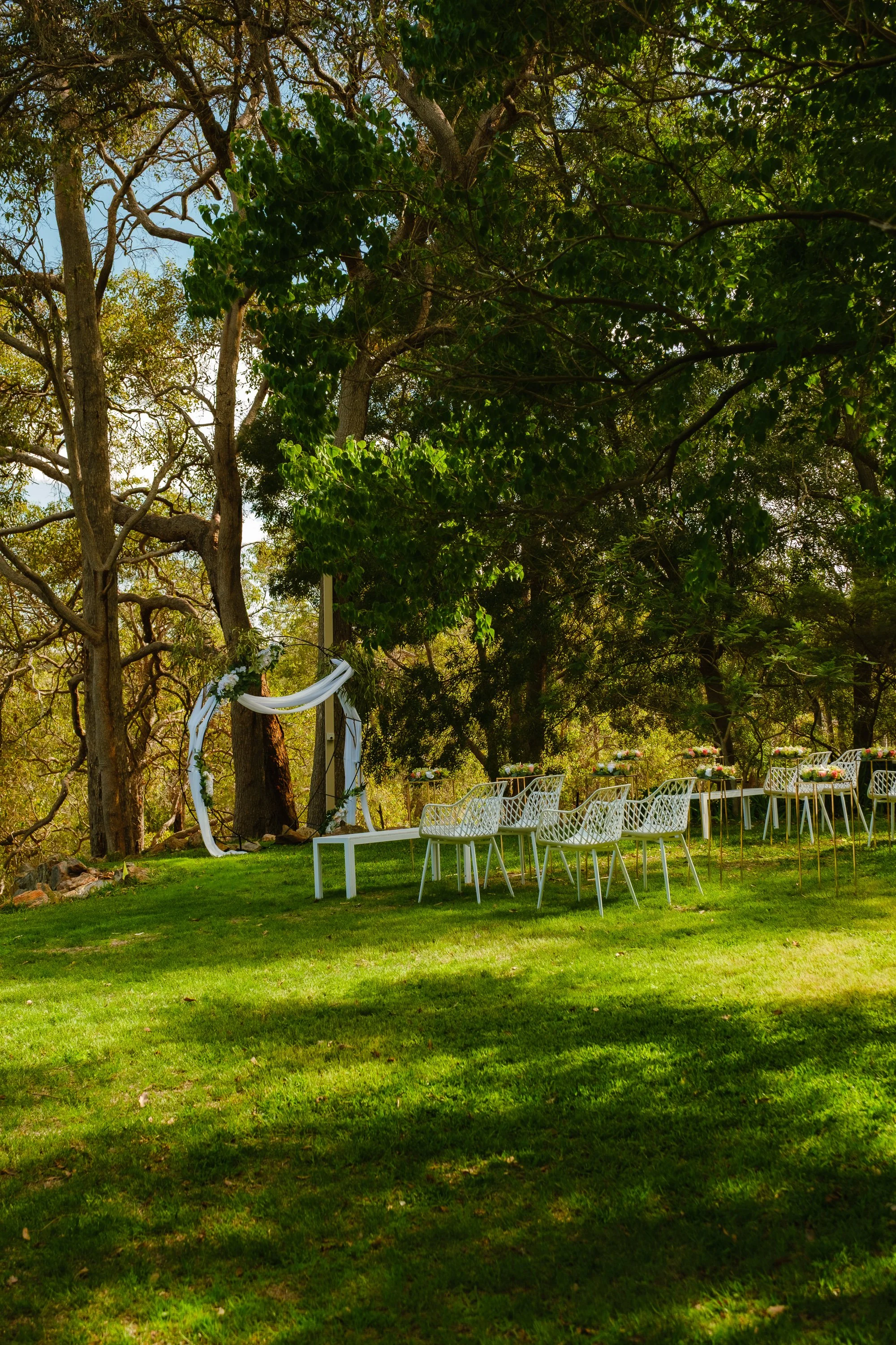 Outdoor wedding setup with white chairs and tables decorated with flowers under large leafy trees.
