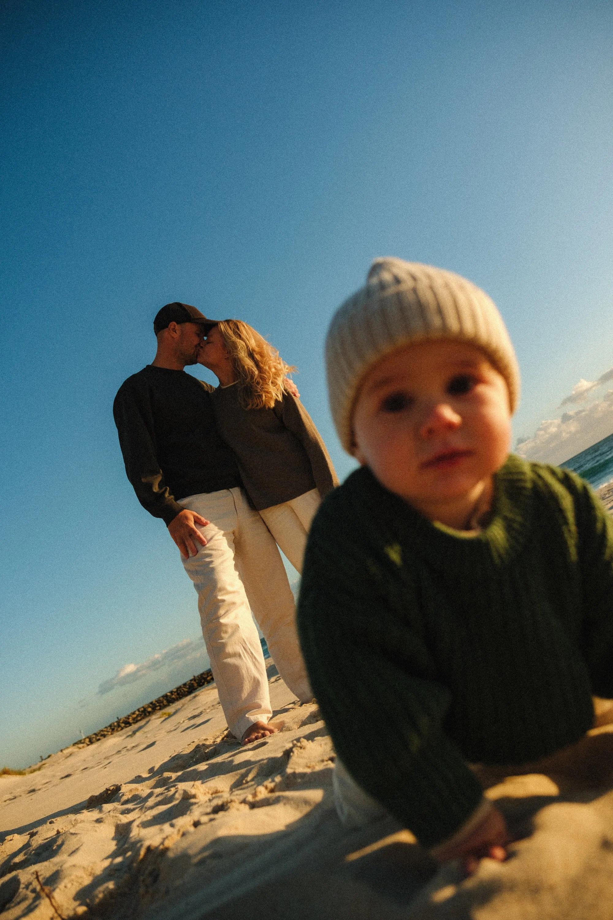 A couple is kissing on the beach during sunset, with a young child in the foreground wearing a knit hat, looking towards the camera.