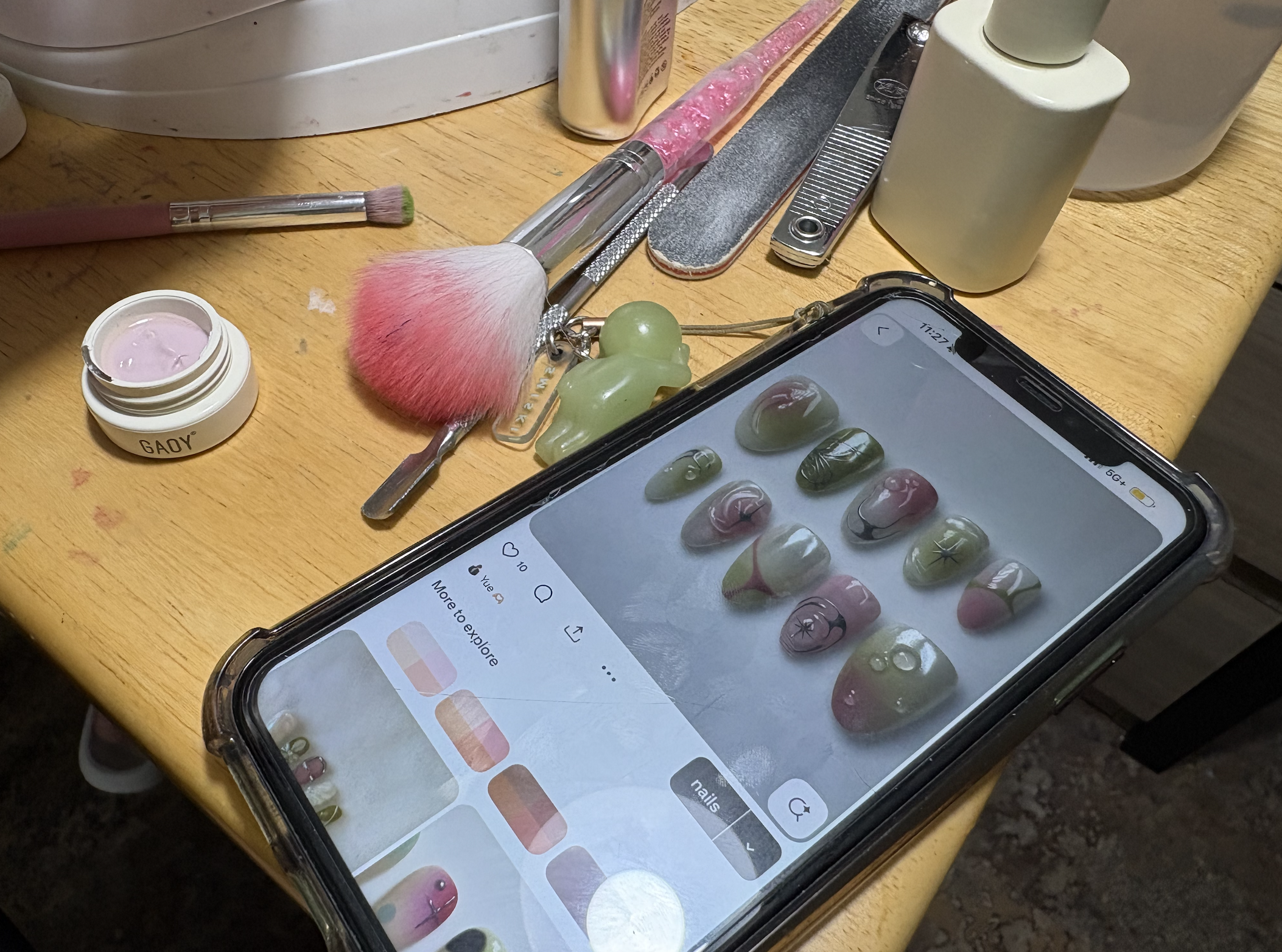 A wooden table holding a variety of manicure tools, including brushes, scissors, and nail files. There's a small open container with pink cream or gel, a smartphone displaying nail art options, and other supplies for nail care.