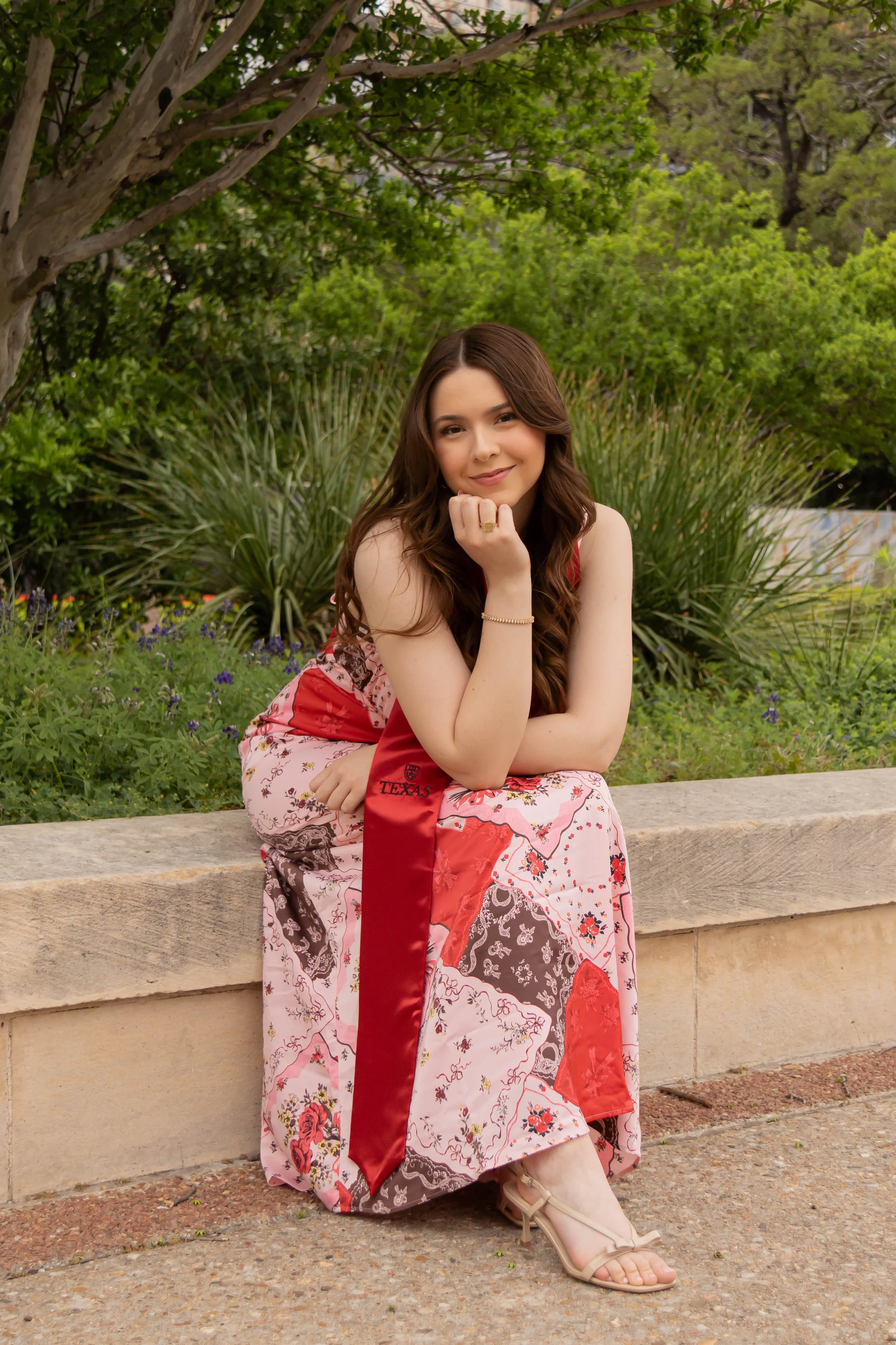 A young woman sitting on a stone bench outdoors, smiling, wearing a floral dress with red accents, and beige high-heeled sandals. Green trees and plants are in the background.