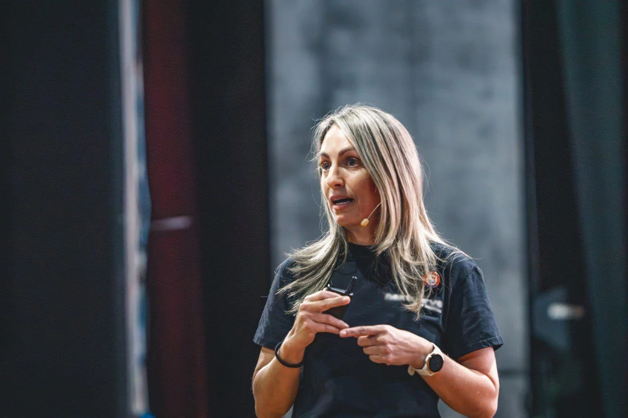 A woman with blonde hair wearing a black shirt and a microphone headset is speaking on stage, holding a small device in her hands.