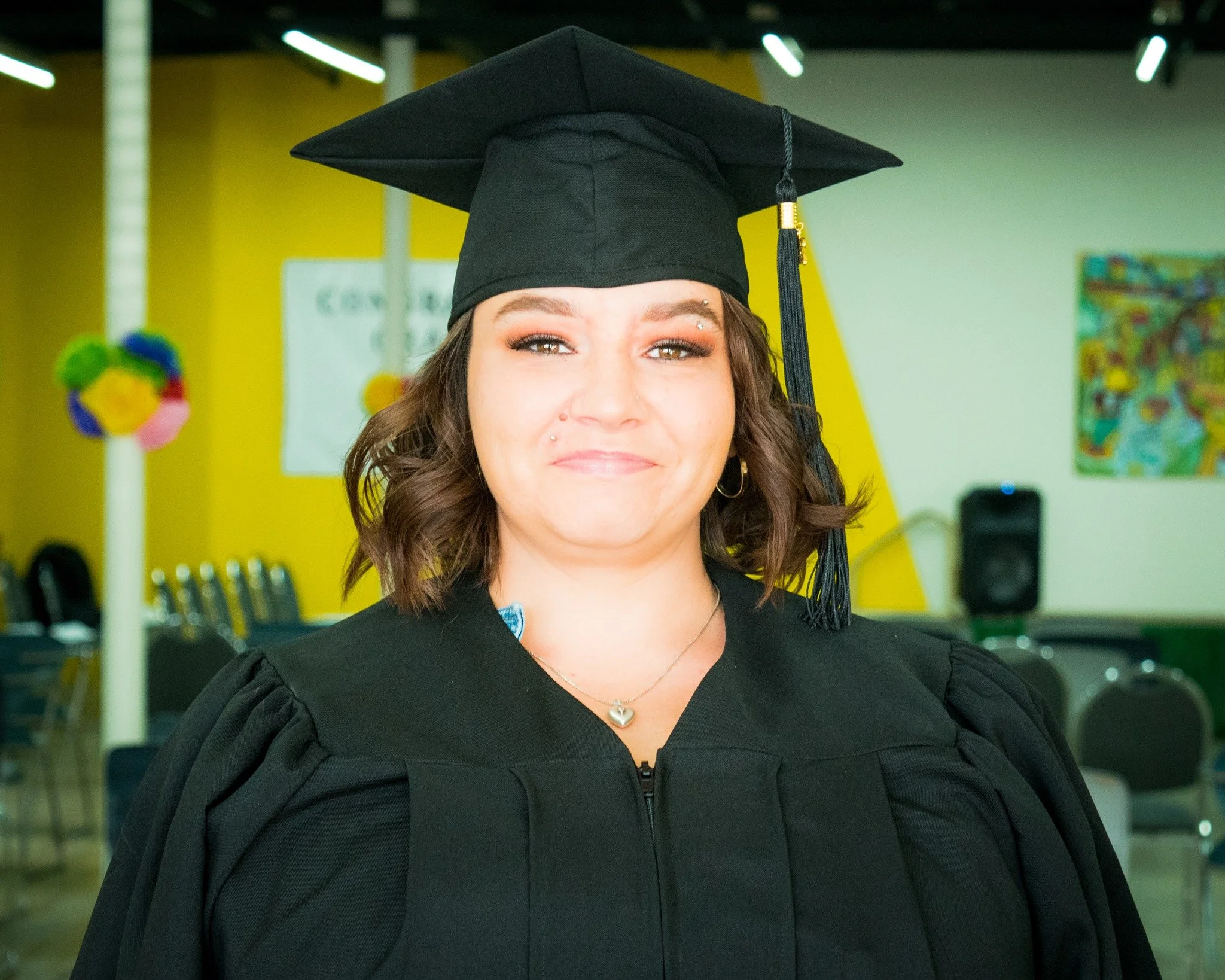 A young woman in a black graduation cap and gown, smiling, inside a decorated hall with yellow walls and colorful artwork.
