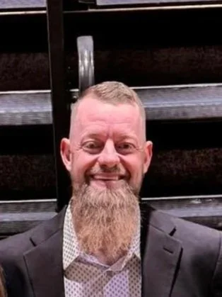 A man with a beard and short hair, wearing a suit jacket and a collared shirt, smiling in front of a staircase.