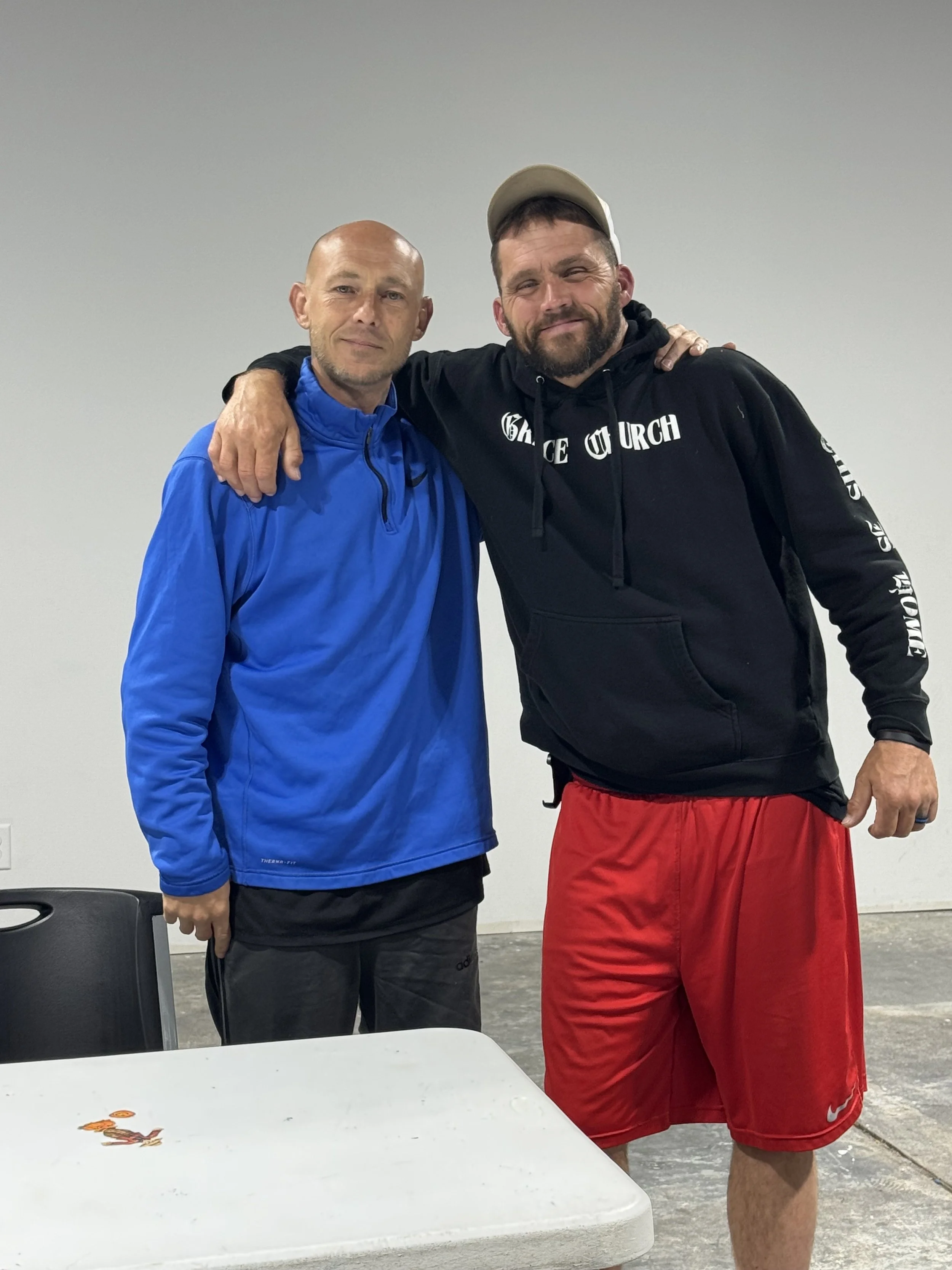 Two men standing together with their arms around each other's shoulders, smiling at the camera. One man is wearing a blue athletic jacket and dark pants, while the other is wearing a black hoodie with white text and red shorts, and a beige cap. They appear to be indoors, possibly in a gym or community center.