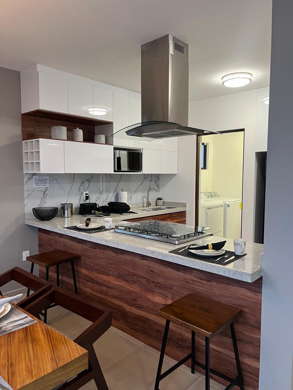 Cocina moderna con barra de granito y taburetes de madera, estufa de gas en la isla, horno microondas, campana extractora de acero inoxidable, gabinetes blancos y detalles en madera, electrodomésticos en la encimera, área de lavandería en el fondo con lavadora y secadora.