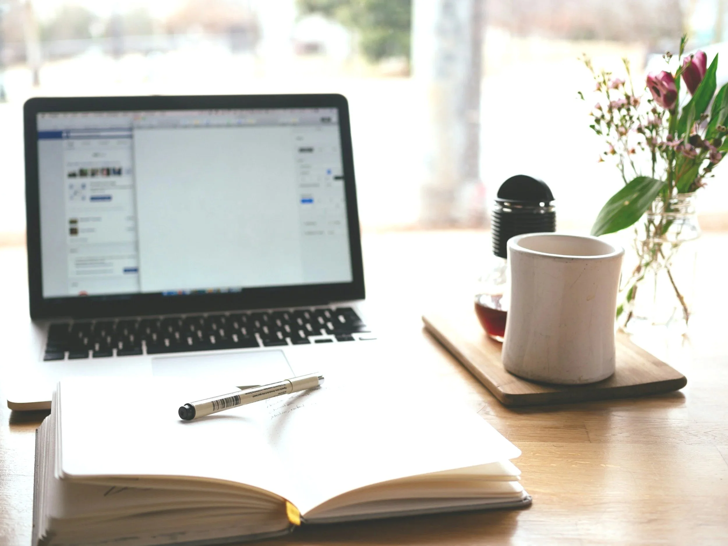 Workspace with a laptop, open notebook with a pen, coffee mug, glass jar with flowers, and a small bottle on a wooden desk near a window
