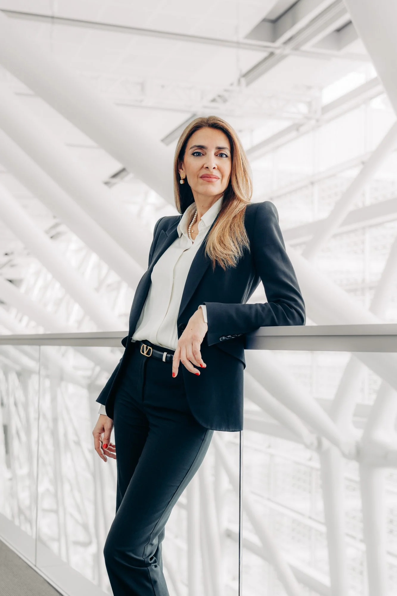 Businesswoman wearing a black blazer and white shirt standing in an airport terminal or modern building with metal beams and glass architecture.