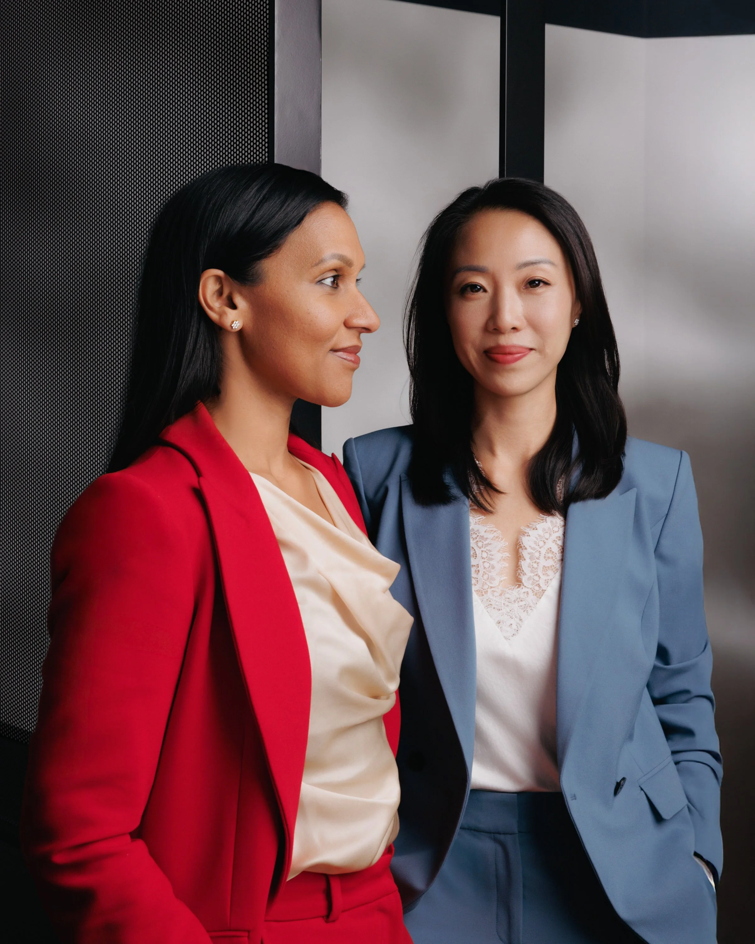 Two women standing close in an elevator, one in a red suit and the other in a blue suit. They are looking at the camera, with the woman in the red suit being slightly in profile.
