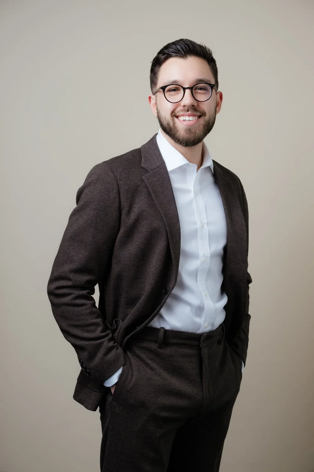 A young man with dark hair, glasses, and a beard, dressed in a white shirt and a dark blazer, smiling and standing against a plain beige background.