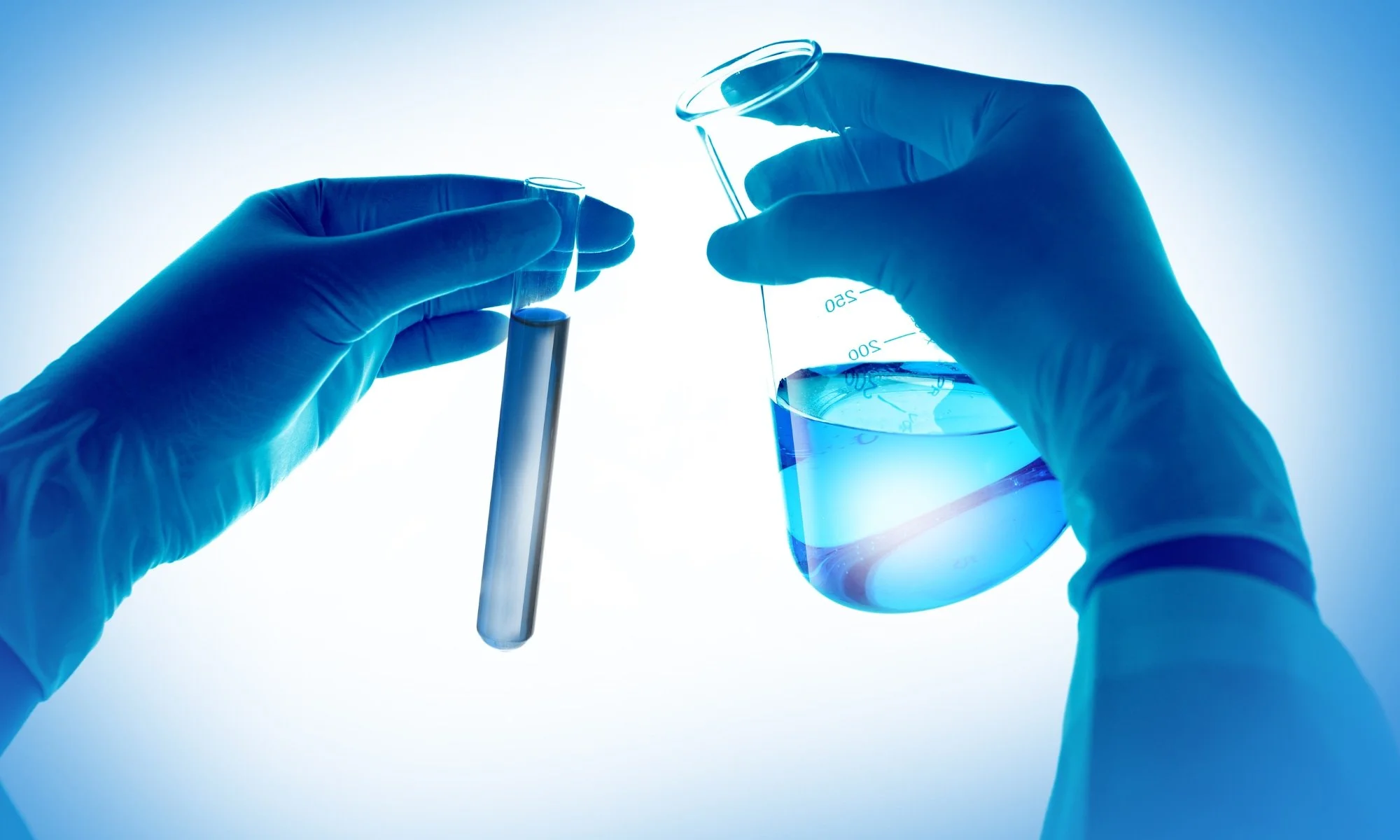 Gloved hands holding laboratory glassware with blue liquid, testing tubes and beaker in a scientific setting.