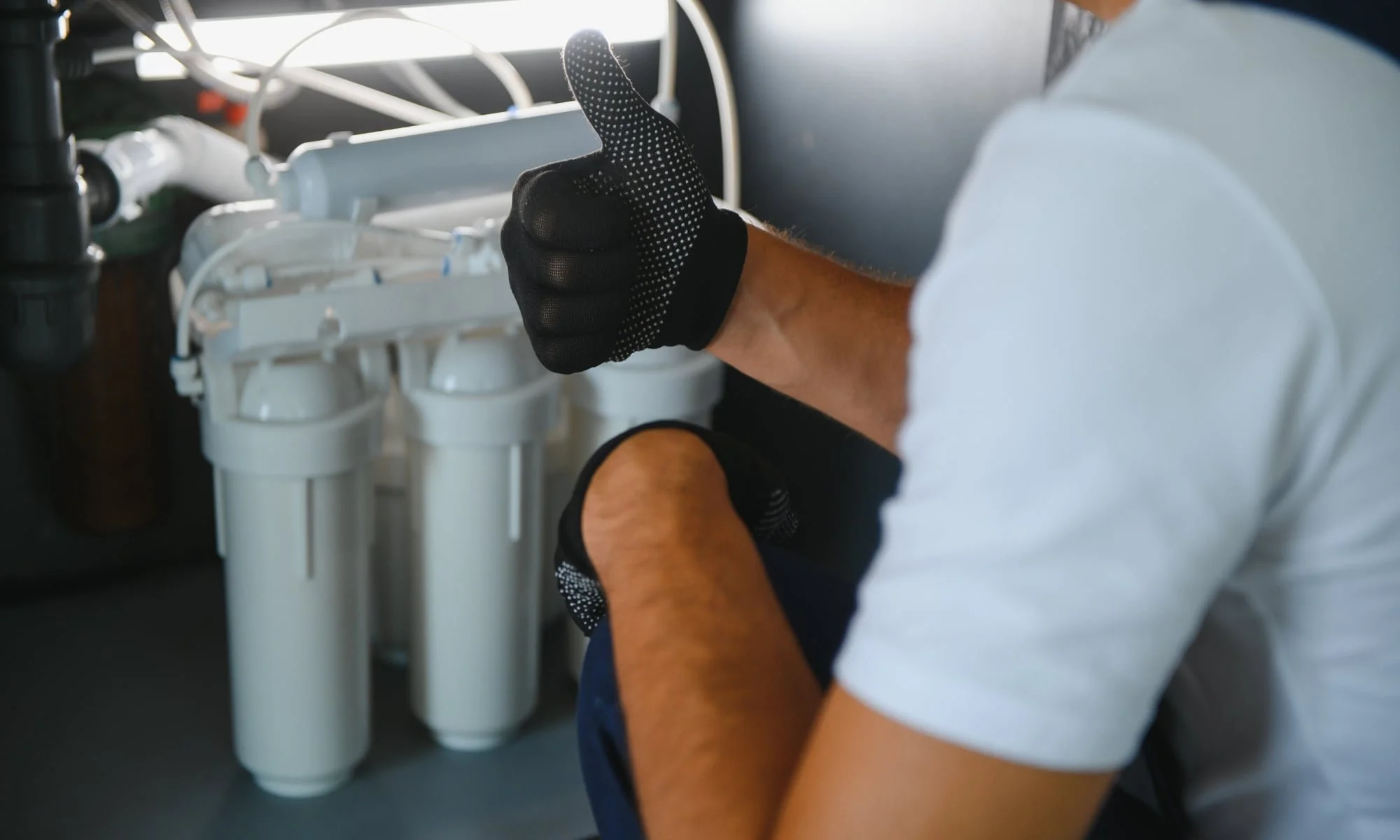 A person wearing black dotted gloves giving a thumbs-up while working on a water filtration system with multiple white filters.