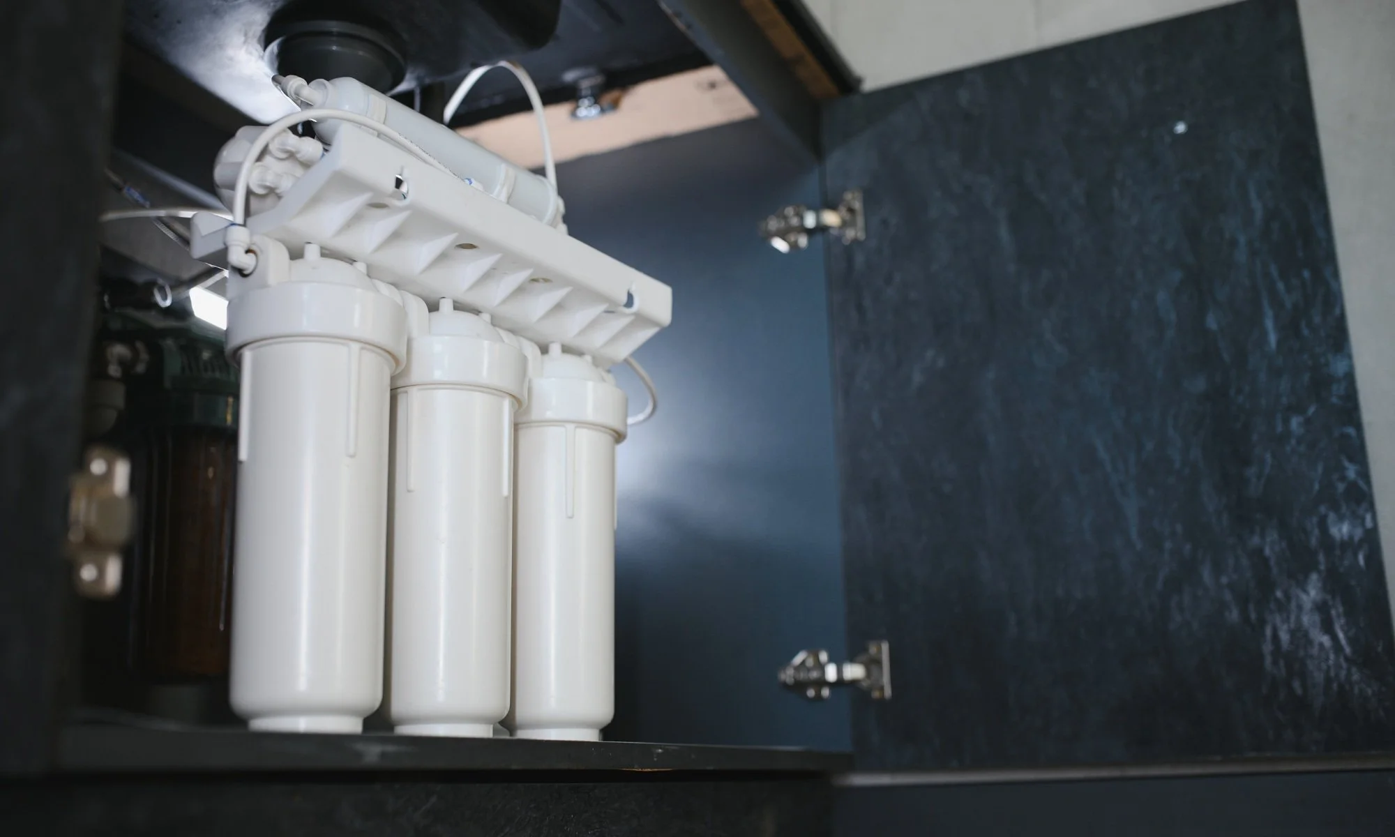 Under-sink water filtration system with white filter cartridges inside a black cabinet.