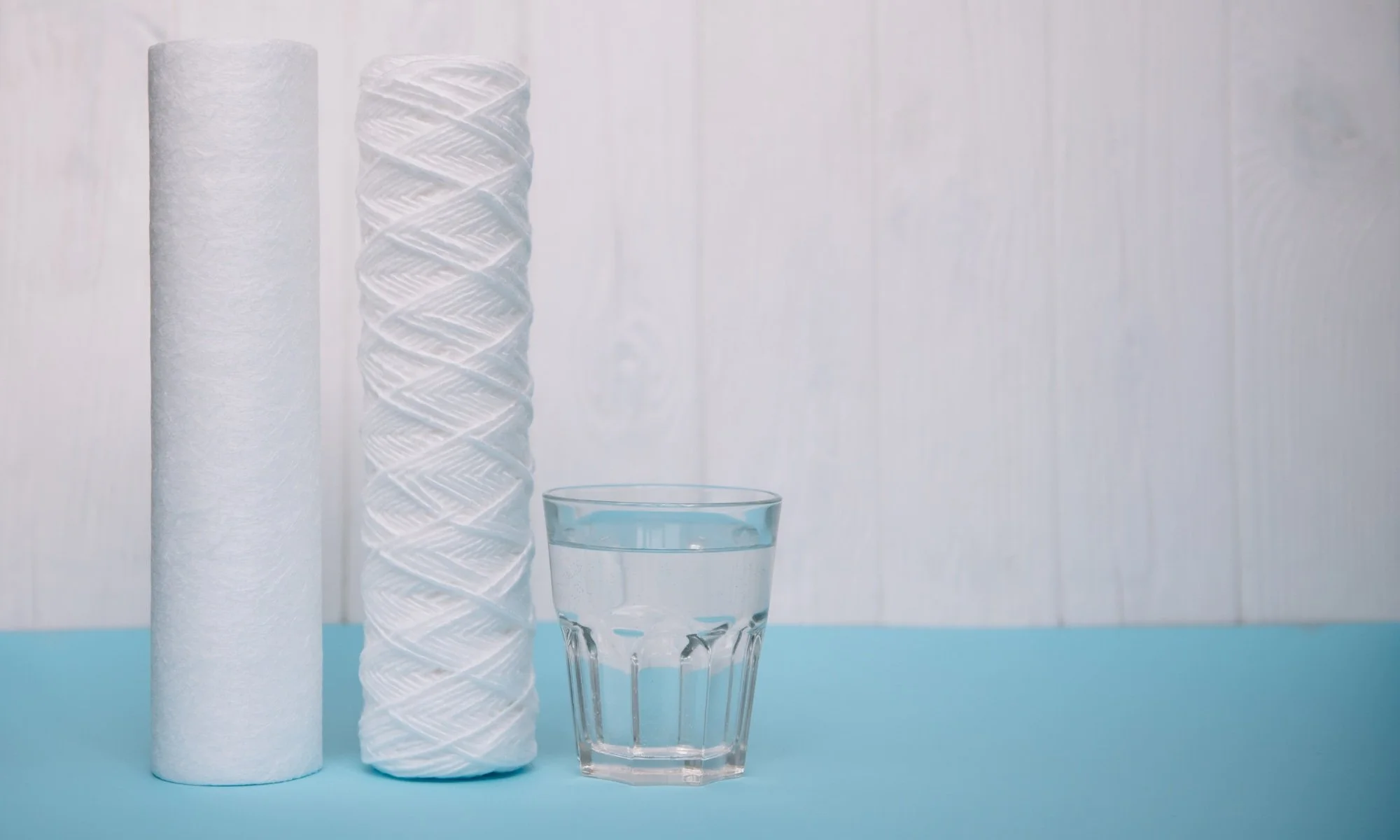 Two water filter cartridges, one plain and one with a patterned surface, standing next to a glass of water on a blue surface with a white wooden background.