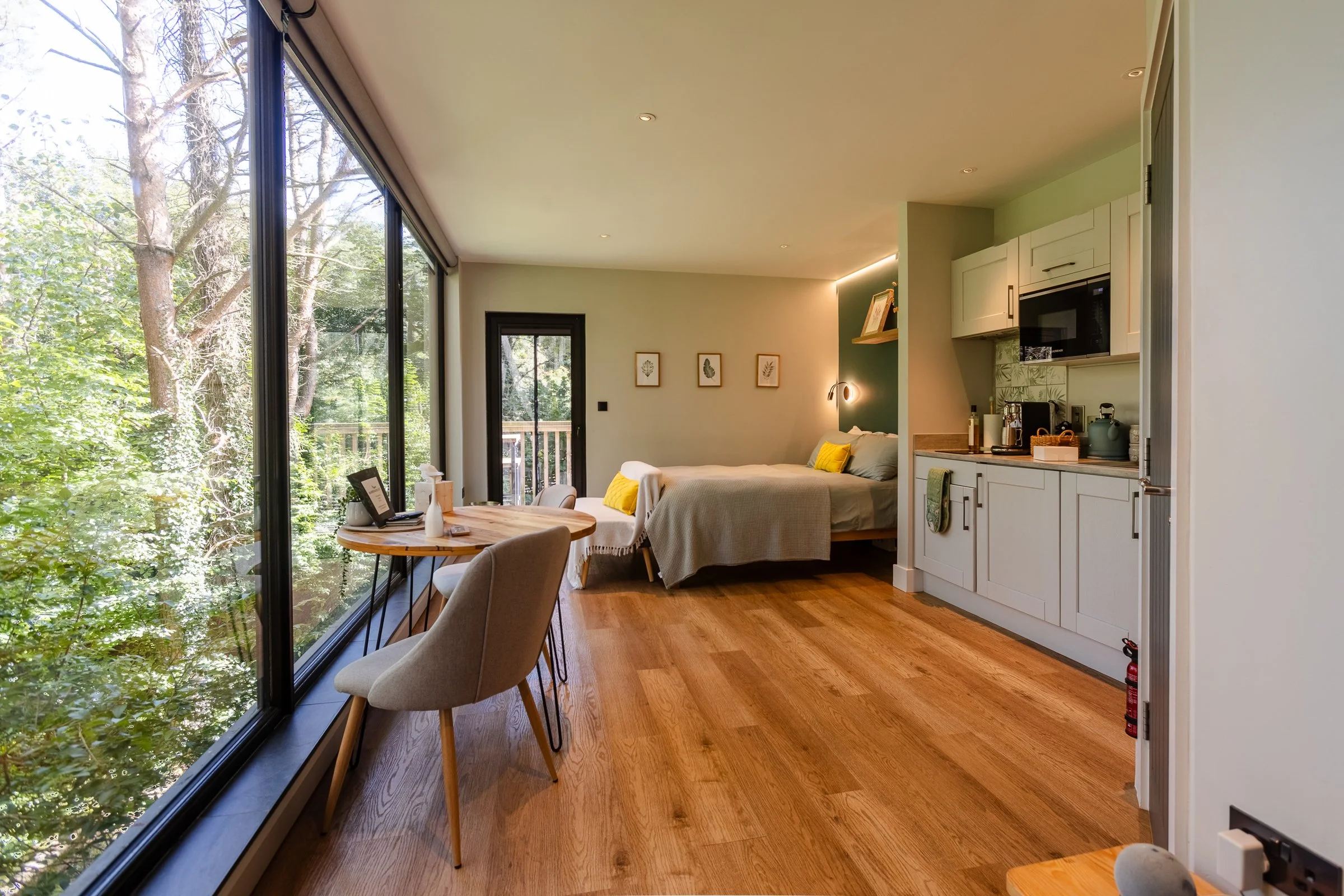 Interior view of a modern studio apartment with a large window showing trees outside, a bed with yellow pillows, a small kitchenette, and a wooden dining table with two chairs.
