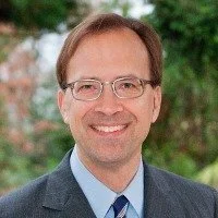 Portrait of a smiling man with glasses, wearing a suit and tie, outdoors with greenery in the background.