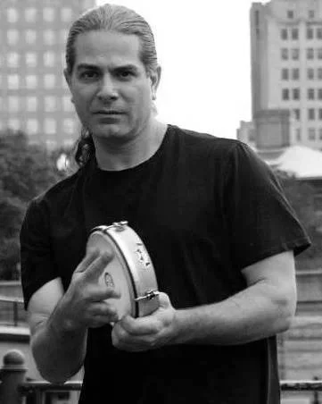 A man with long hair holding a tambourine in an urban setting with tall buildings in the background.