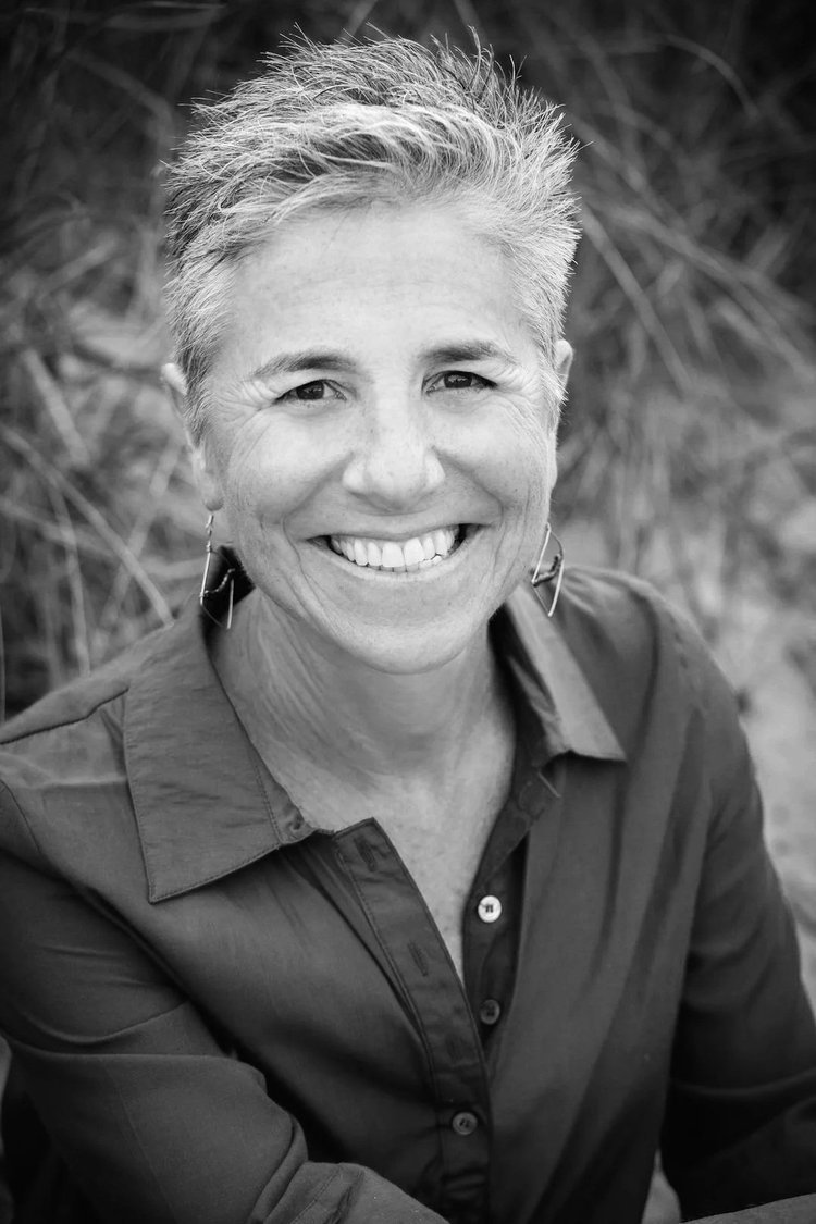 Black and white portrait of a smiling woman with short, spiky hair, wearing earrings and a button-up shirt, outdoors with blurred foliage in the background.