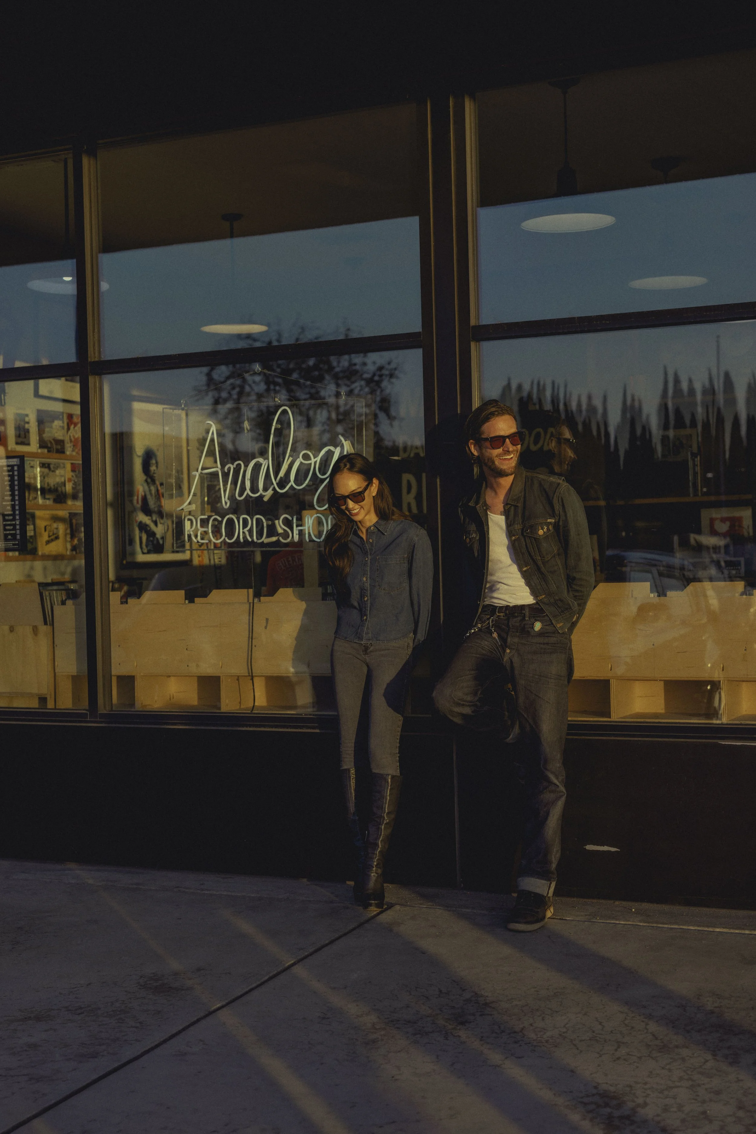Two people, a woman and a man, standing outside of a record store with a large window reflecting the sky. The store has a neon sign that says 'Analogue Record Shop'. The woman is wearing a denim jacket, jeans, and sunglasses, while the man is wearing