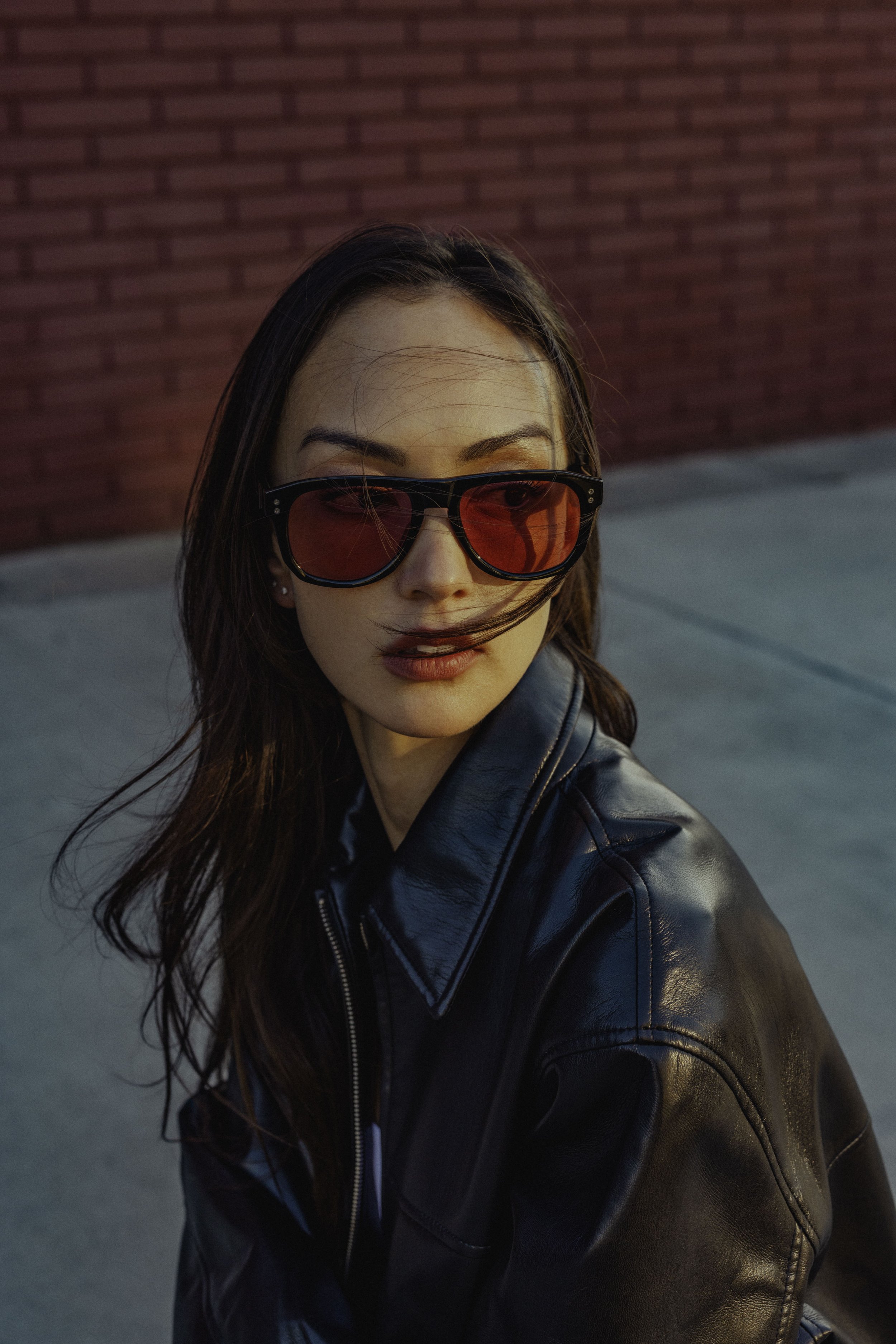 A woman with brown hair wearing red-tinted sunglasses and a black leather jacket. She is outdoors with a red brick wall in the background.