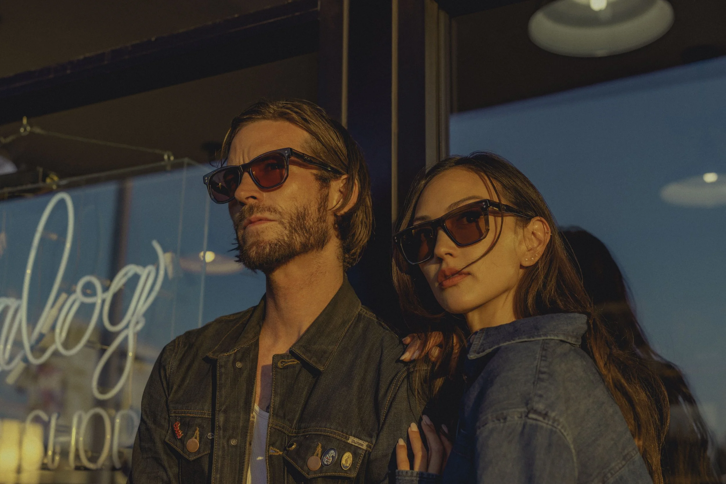 A man and woman wearing sunglasses standing close together outside at sunset, with a neon sign behind them.