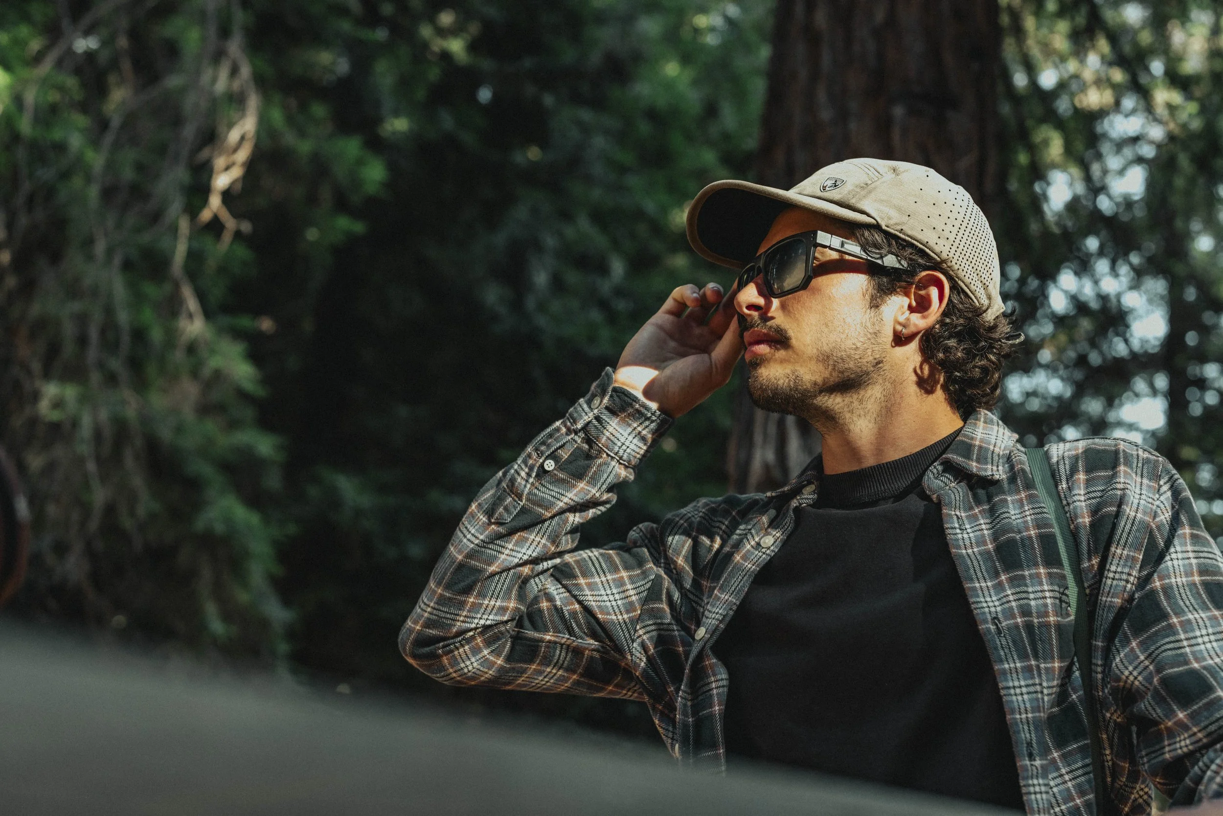 A man with curly hair and a beard, wearing a beige cap, sunglasses, and a plaid shirt, adjusting his glasses while outdoors in a forest.