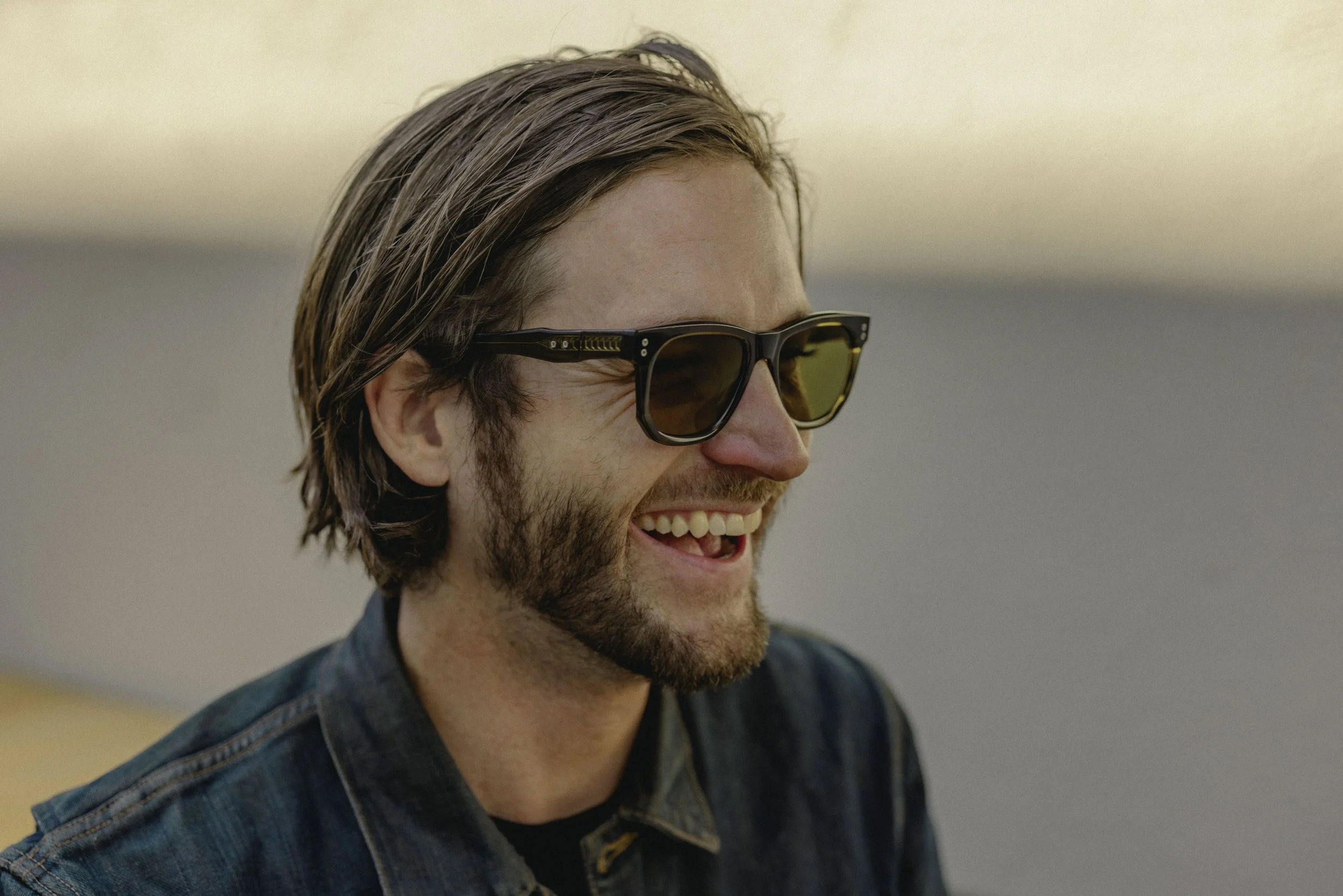 A man with shoulder-length brown hair, wearing sunglasses and a checkered shirt, smiling and looking to the right.