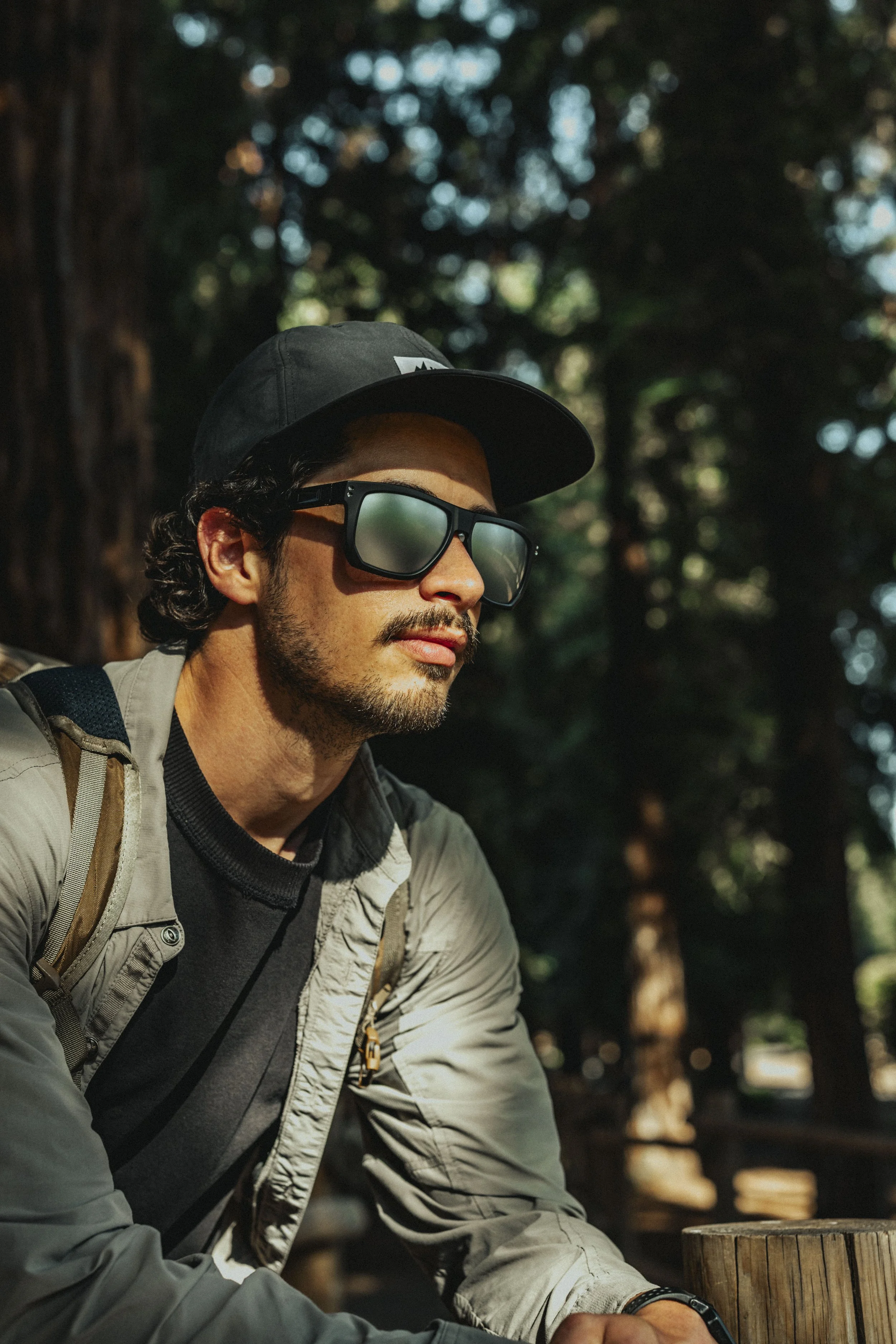 A man with dark, curly hair, wearing sunglasses, a black baseball cap, a black T-shirt, a light-colored jacket, and a backpack, sitting outdoors in a wooded area with trees and sunlight filtering through the leaves.