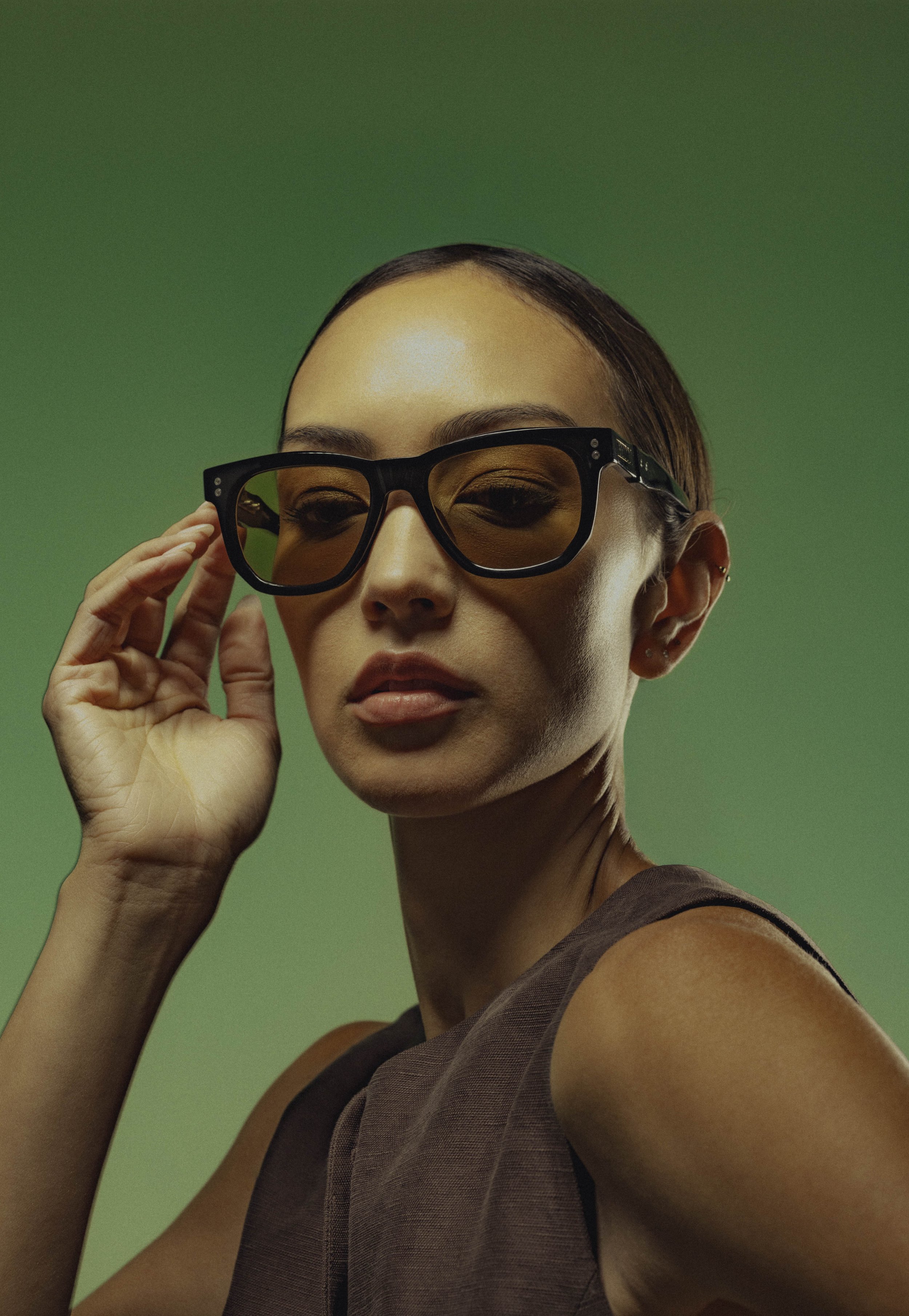 A woman with short brown hair wearing fashion glasses, touching her temple with her left hand, against a green background.