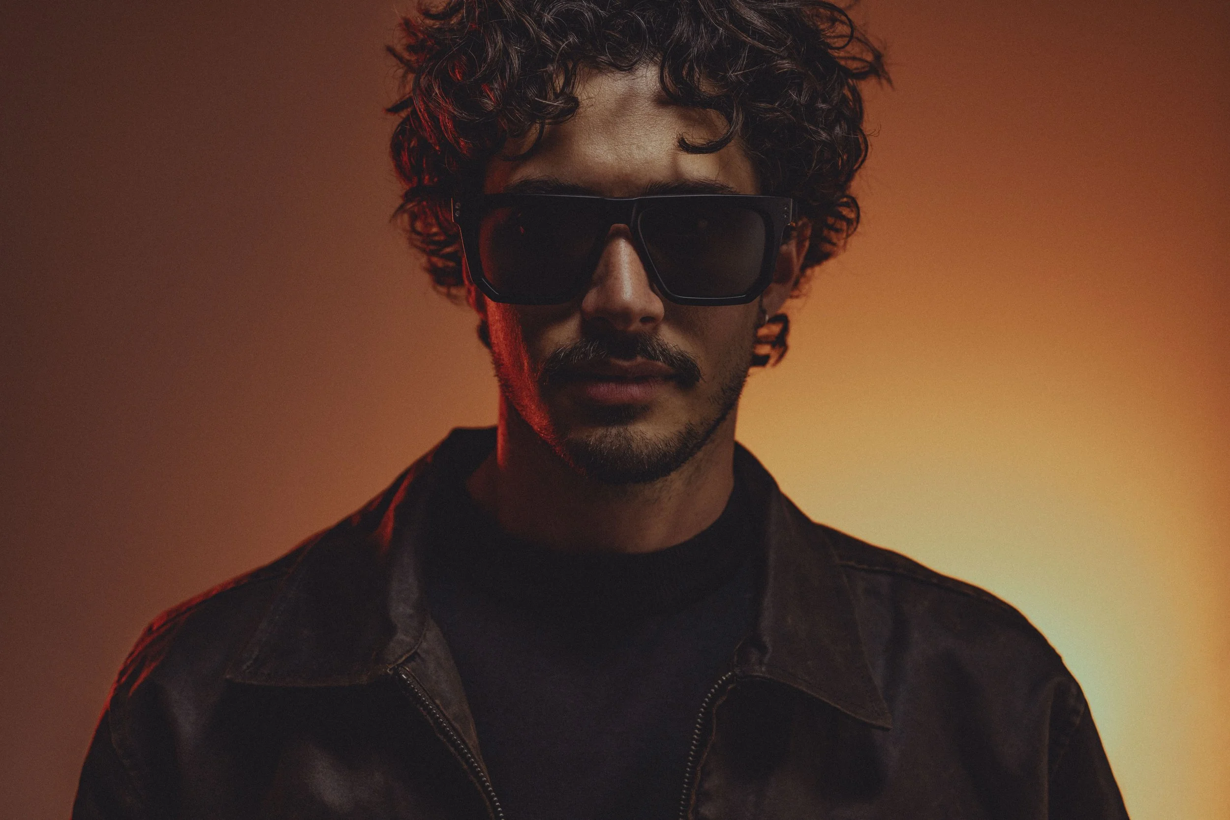 A man with curly hair wearing dark sunglasses and a leather jacket, standing against a gradient orange background.