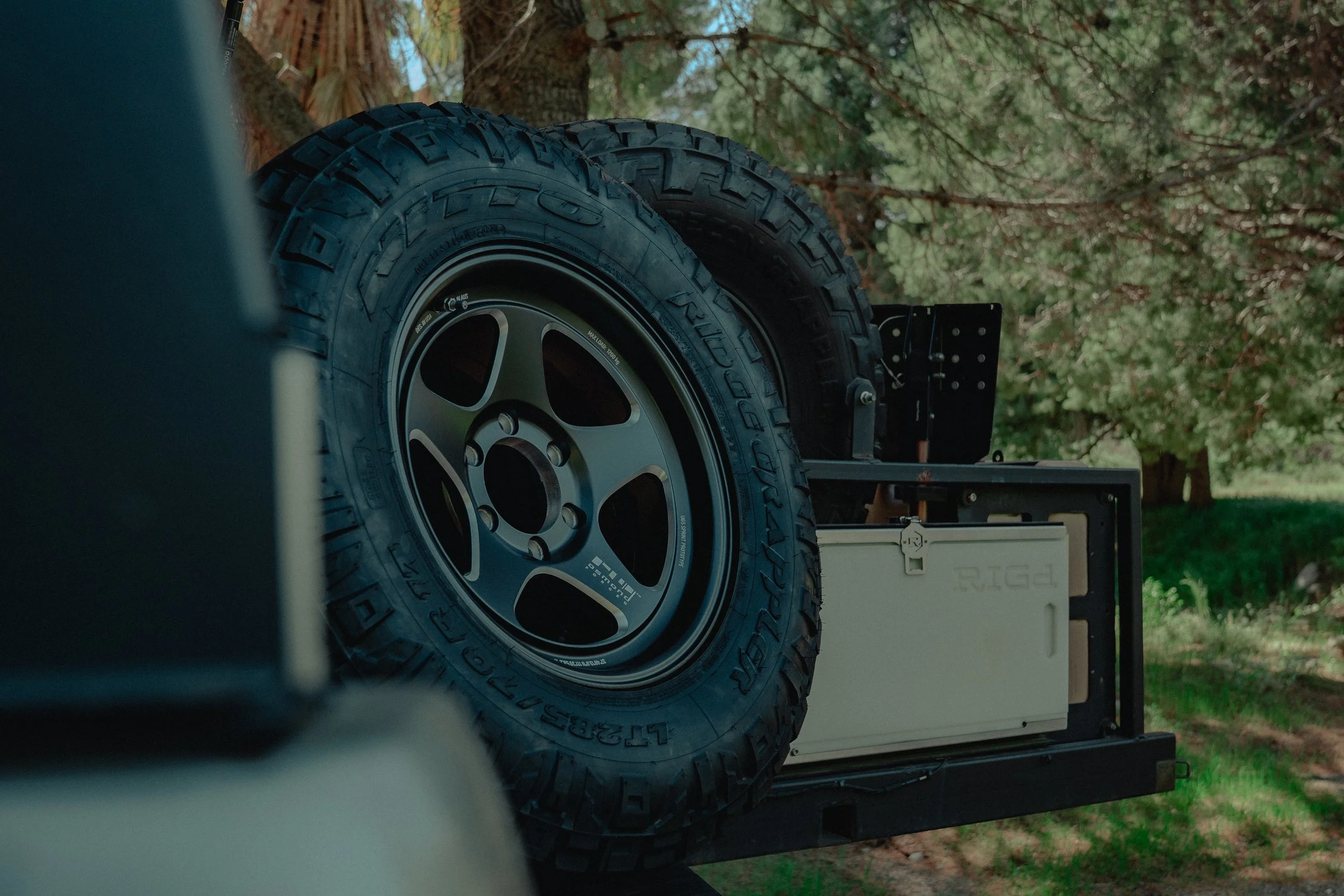 Close-up of off-road tires mounted on a vehicle, with a wooded background and a white container or equipment at the back.