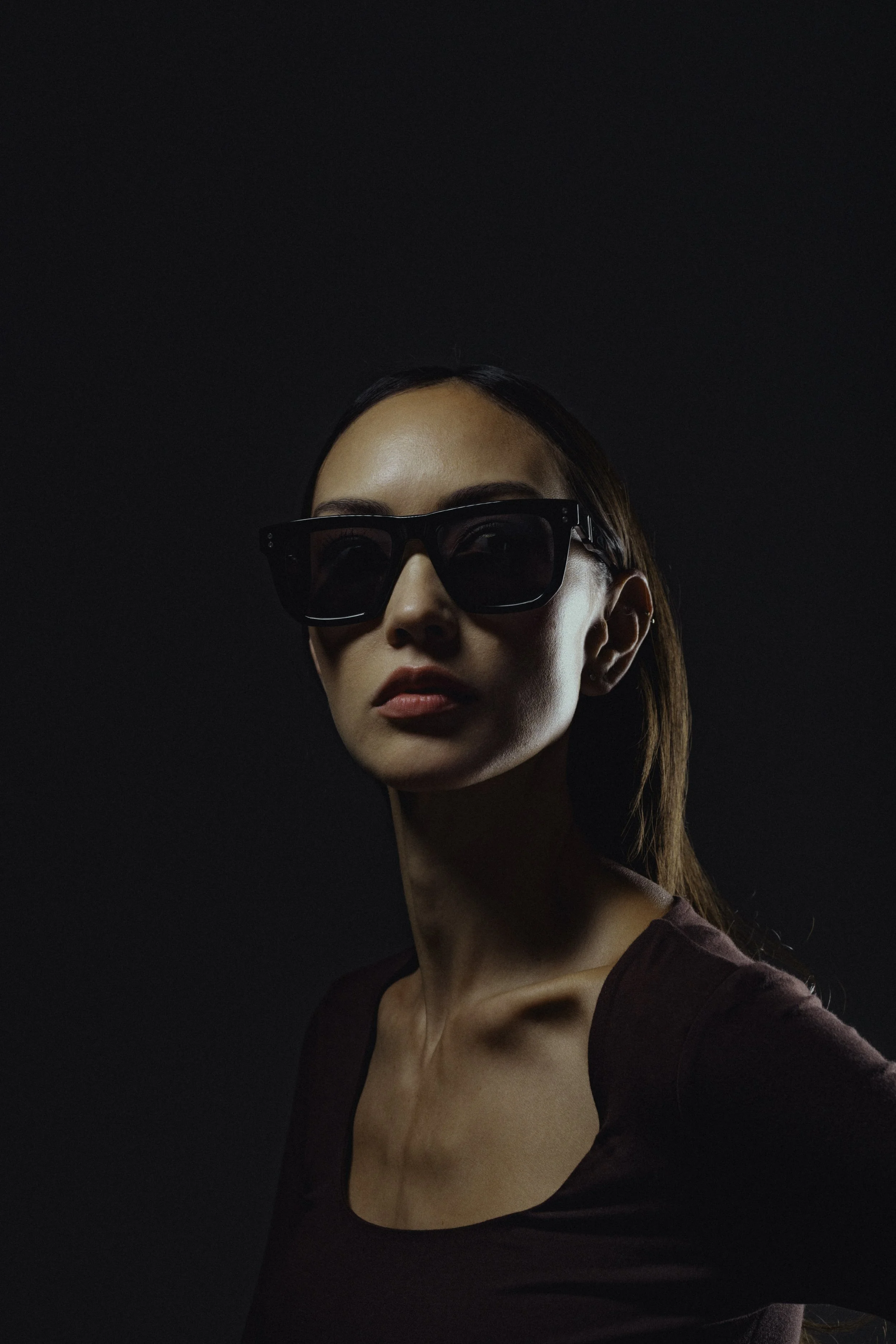 A woman with dark hair and fair skin wearing black sunglasses and a dark purple top standing against a black background.