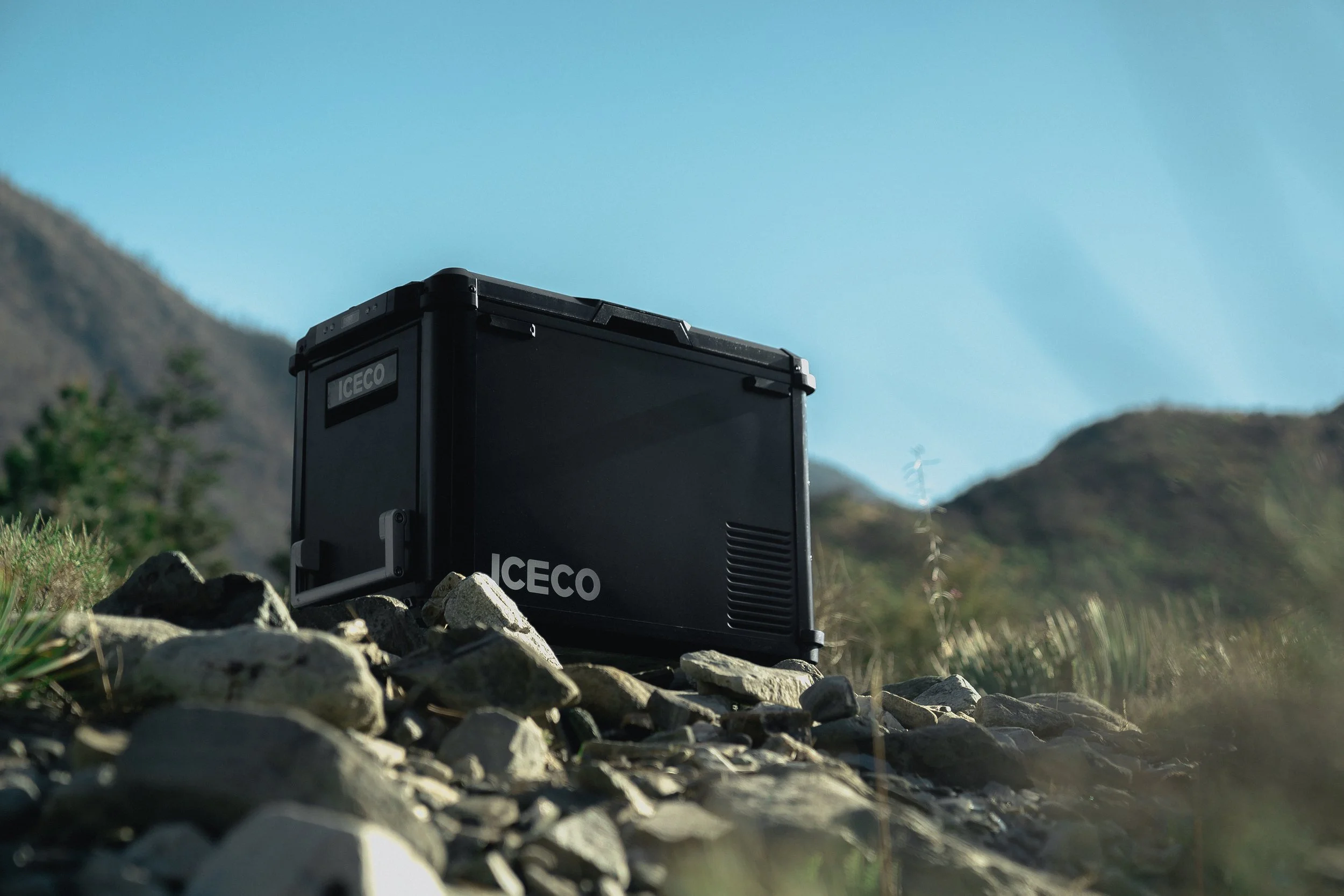 A black ICECO portable refrigerator or cooler placed outdoors on rocky ground with mountains and blue sky in the background.