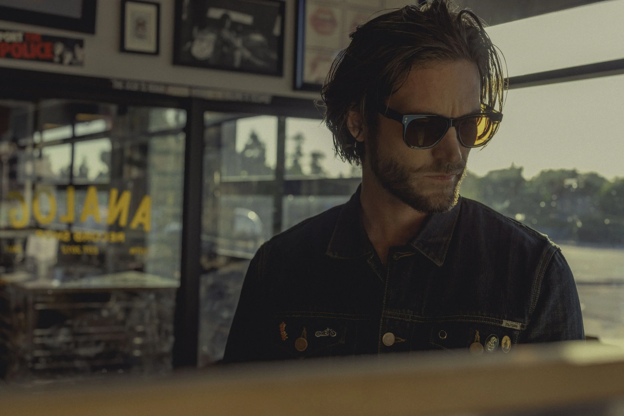 A man with brown hair, beard, and sunglasses looking down inside a diner or cafe. The background shows framed pictures and a window with daylight outside.