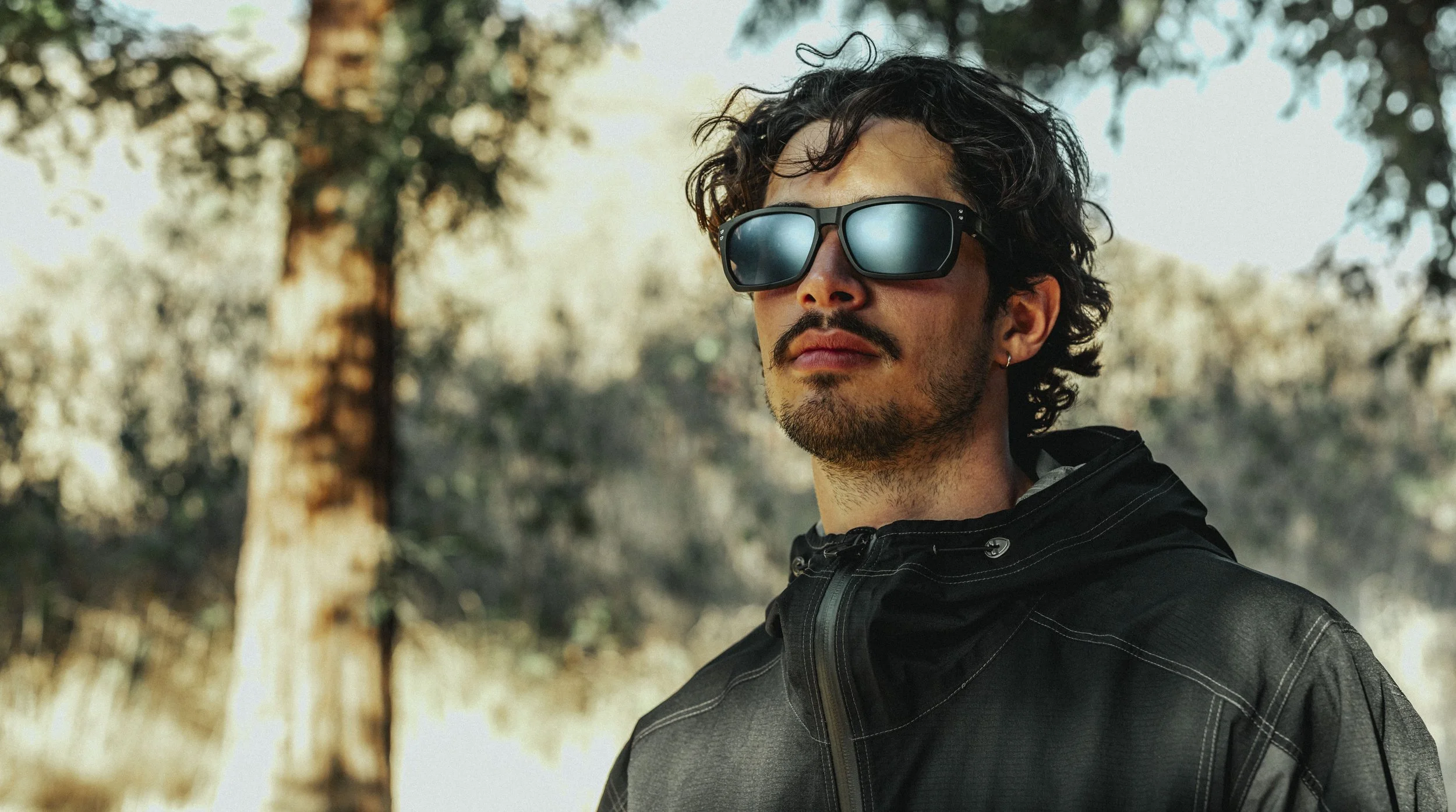 A man with curly dark hair, wearing sunglasses and a black jacket, standing outdoors among trees.