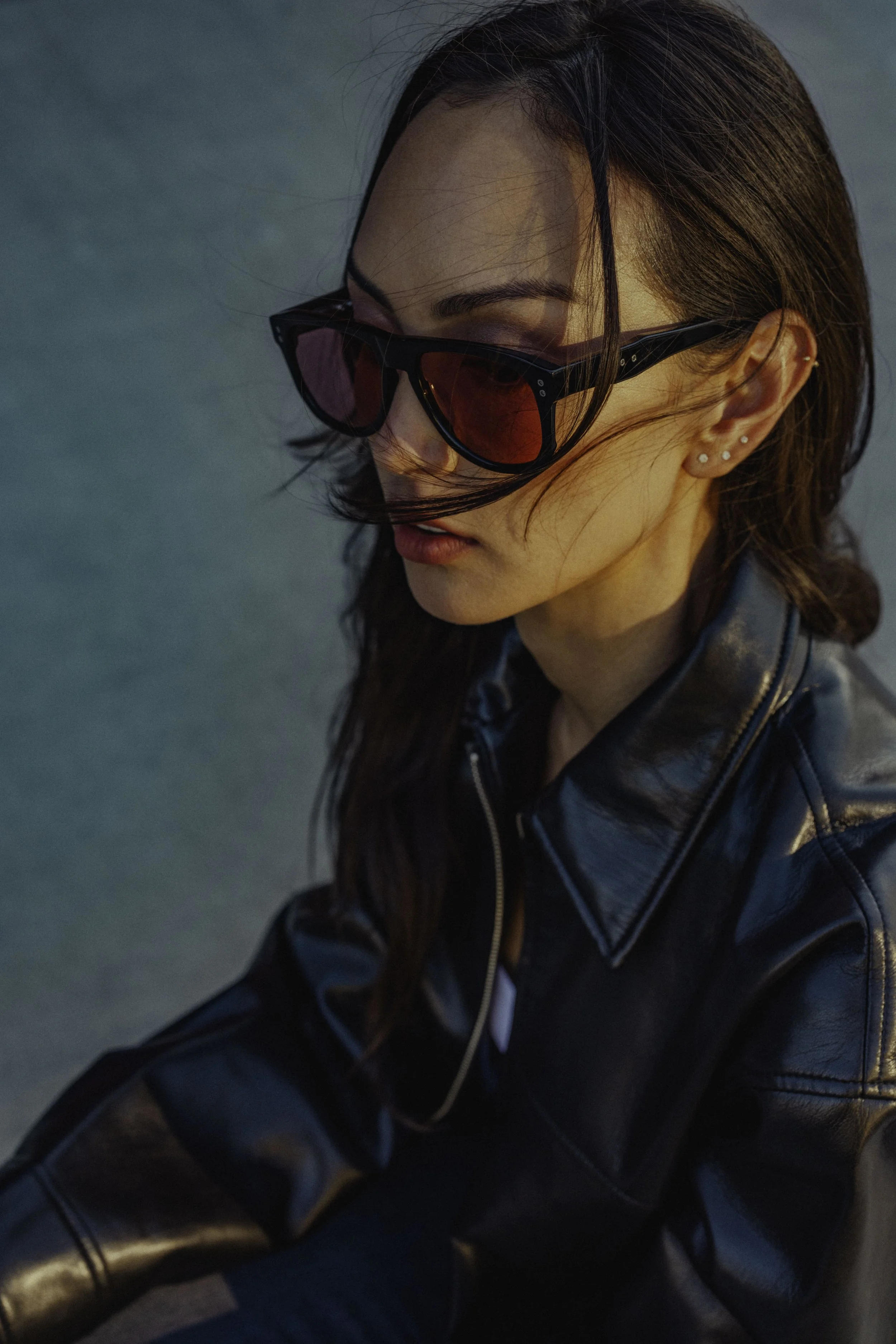 Close-up of a woman wearing black sunglasses with red-tinted lenses, a black leather jacket, and multiple earrings, with her hair partially covering her face.
