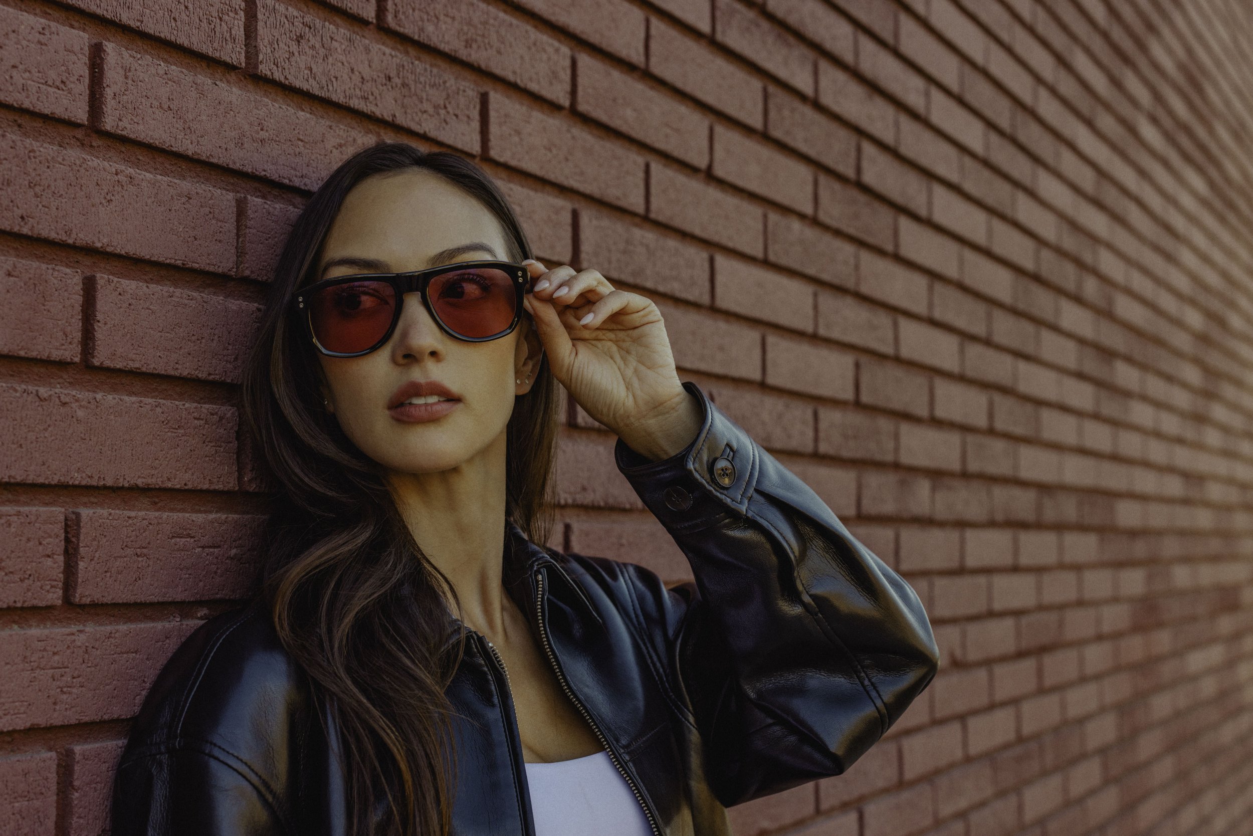 A young woman with long dark hair wearing red-tinted sunglasses, a black leather jacket, and a white top standing against a red brick wall, slightly turning her head while adjusting her sunglasses.