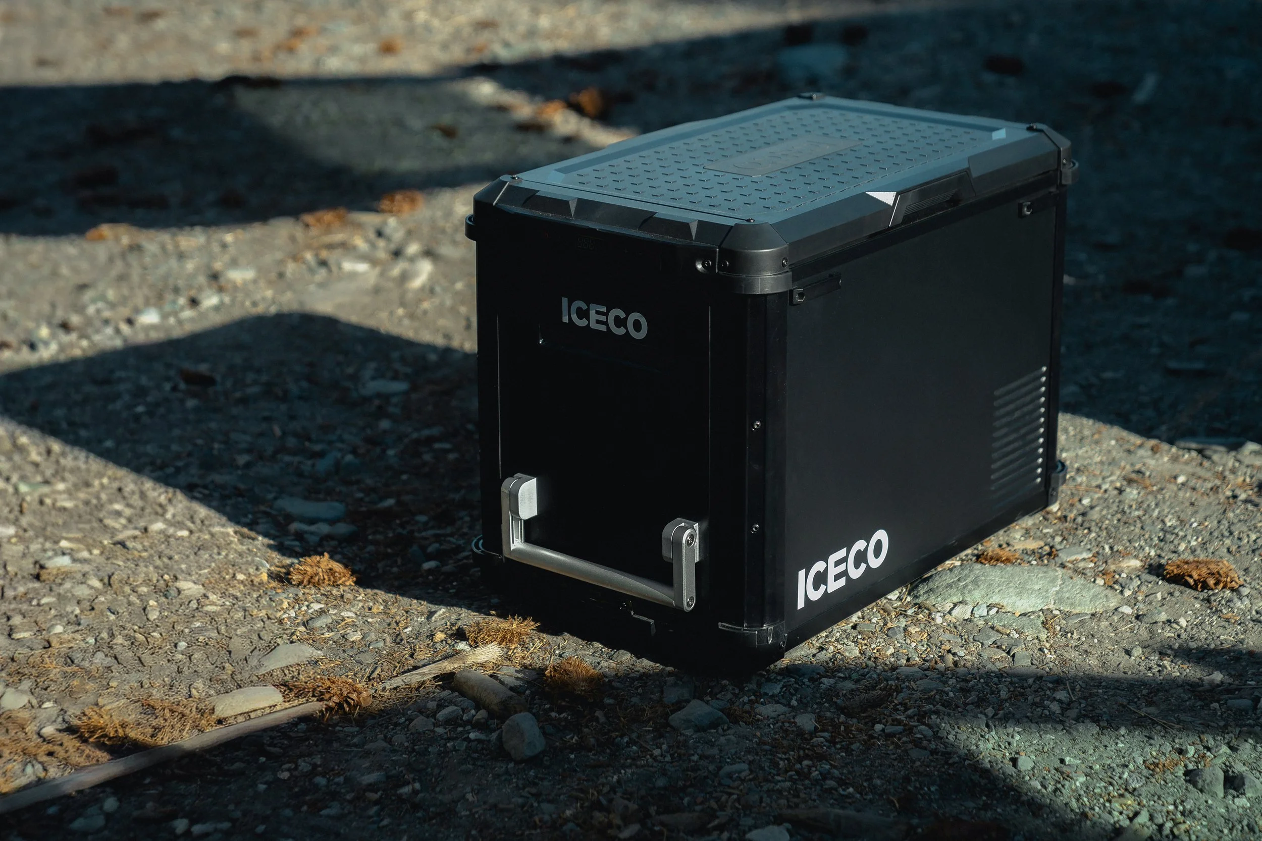 A black portable refrigerator or cooler with the brand name 'ICECO' on it, placed on a gravel surface outdoors, partially in shadow.