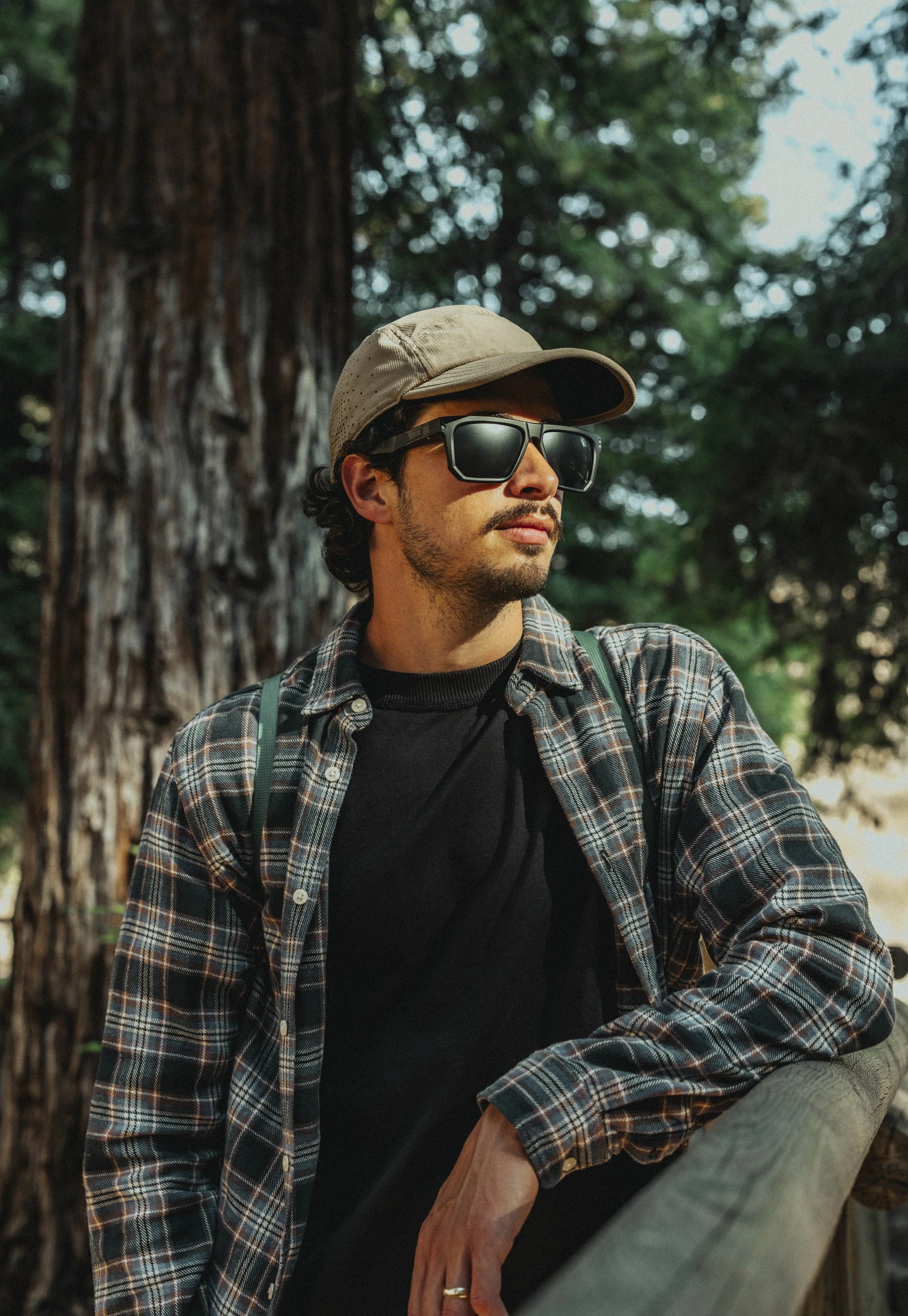 A man wearing sunglasses, a beige cap, a black shirt, and a plaid jacket, leaning on a wooden railing by a large tree in a forest.