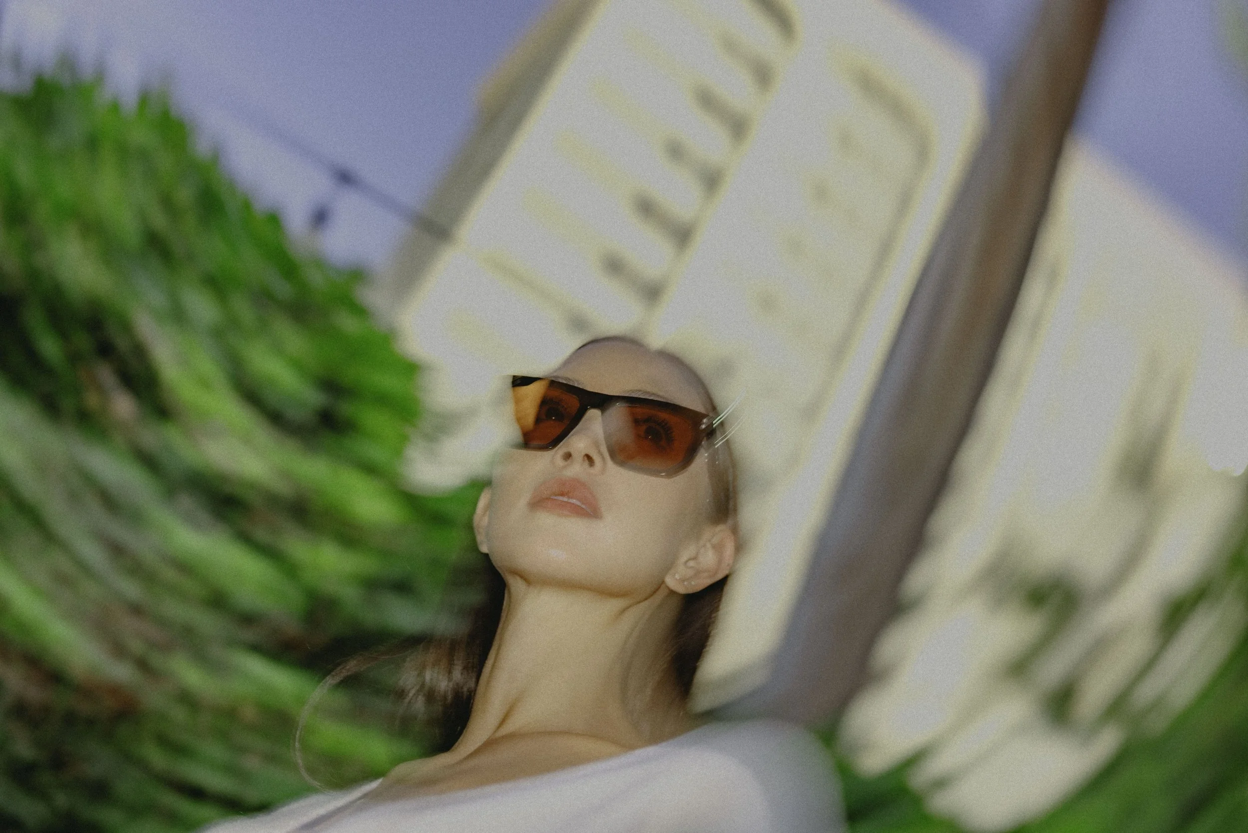 A woman with light skin and red hair wearing sunglasses lying on her back in a bed or on a surface, surrounded by green plants and pillows with blue and beige curtains or blinds in the background.