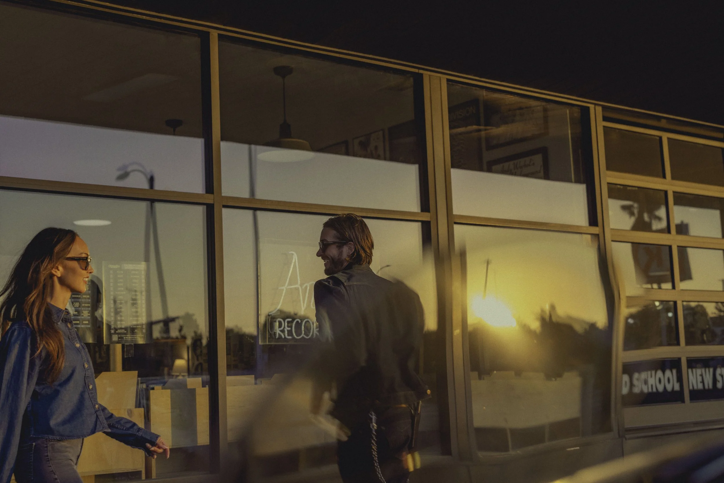 Three people walking past a glass storefront reflecting a sunset sky with orange and yellow hues, and their shadows cast on the glass.