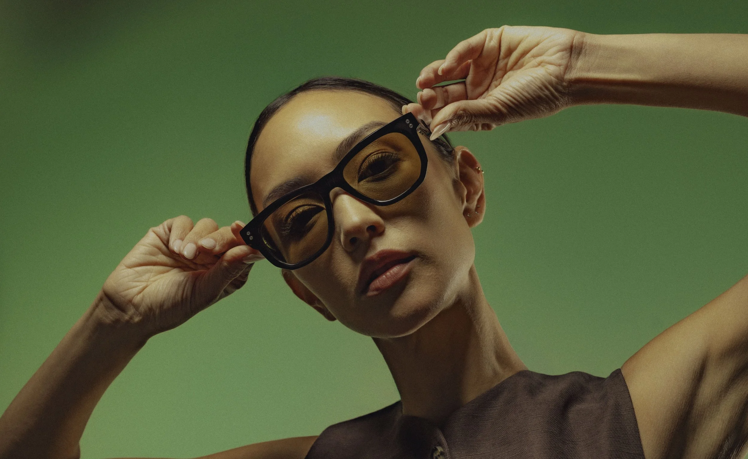 Close-up of a woman with dark hair and glasses adjusting her glasses against a green background.