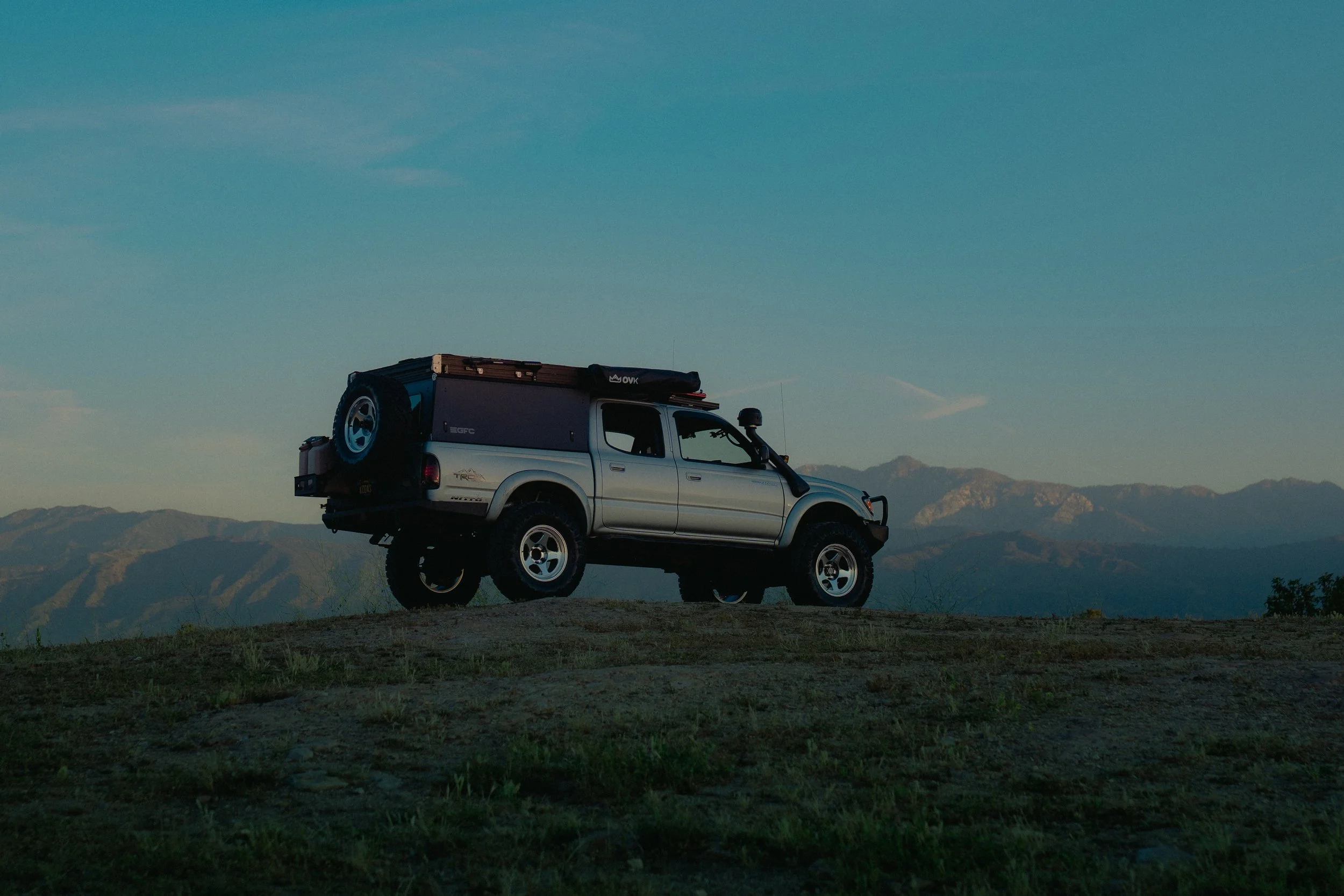 A white off-road vehicle with a roof rack and spare tire parked on a hilltop, with mountain ranges visible in the background and a clear blue sky.