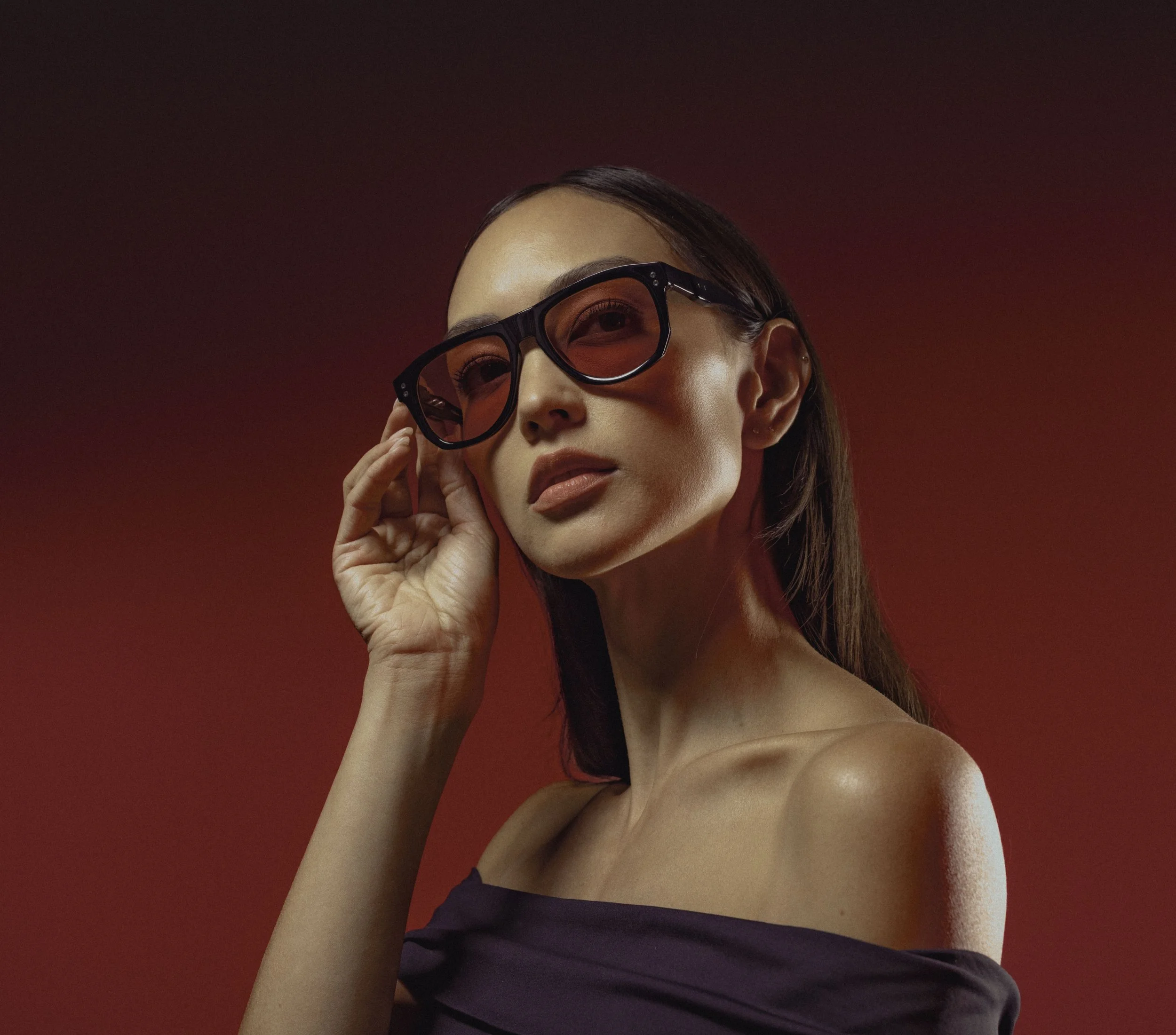 A woman with long brown hair wearing black sunglasses with red tint, lightly touching the side of the sunglasses, against a red background.