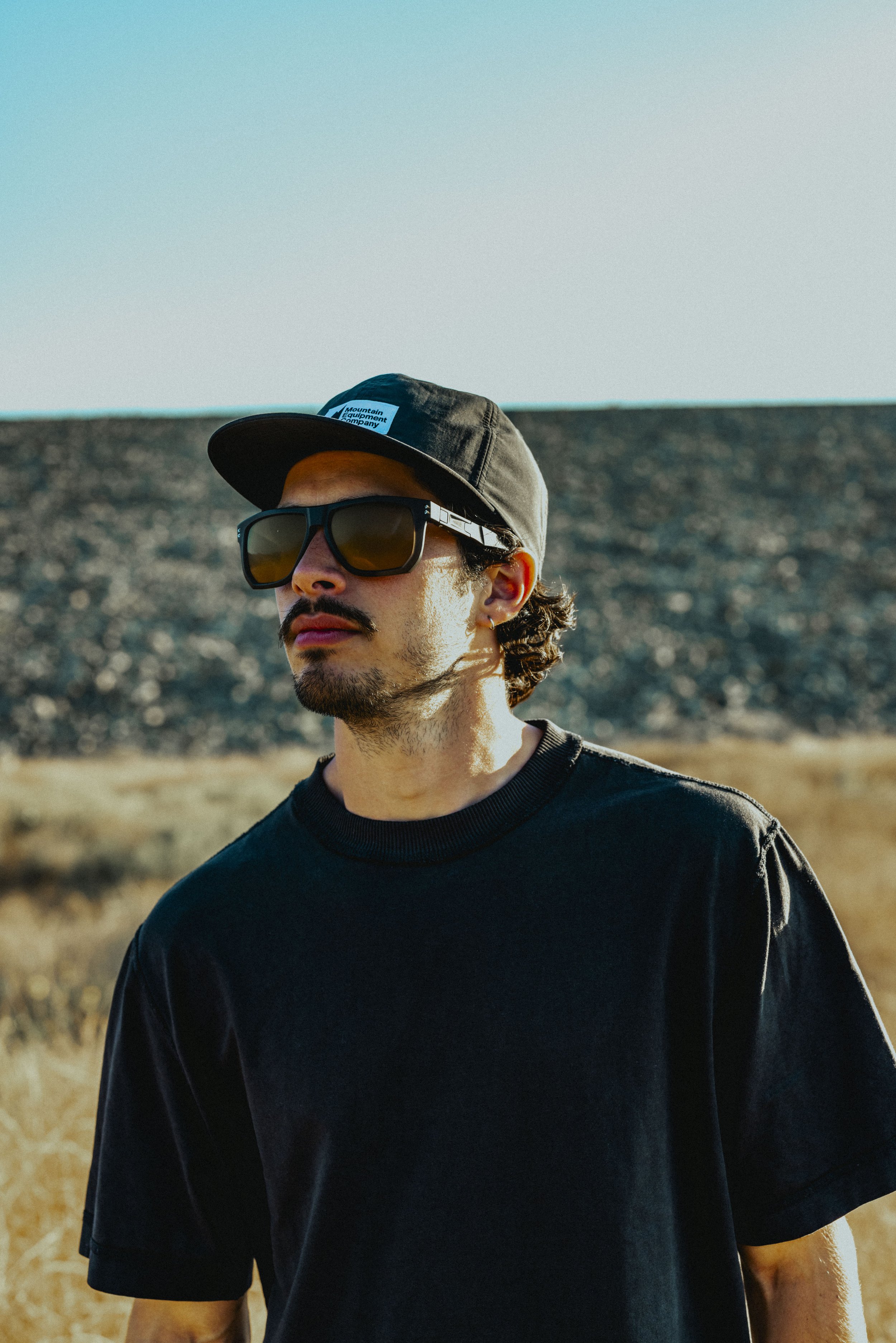 A man wearing a black baseball cap with a blue logo, large black sunglasses, and a black t-shirt, standing outdoors near a dry, hilly landscape under clear sky.