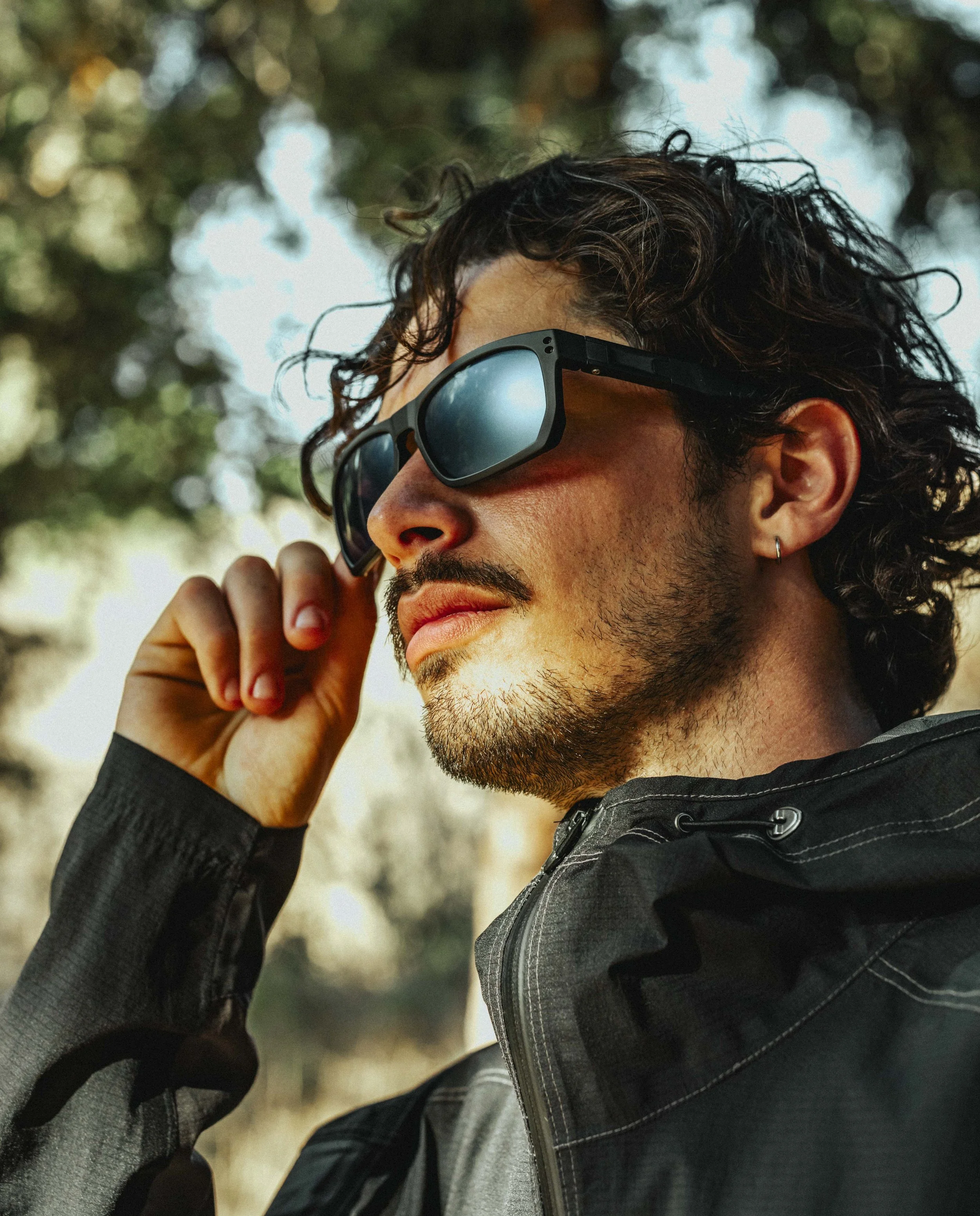 Close-up of a man with curly dark hair, wearing sunglasses and a dark jacket, holding a phone to his ear outdoors with blurred trees in the background.