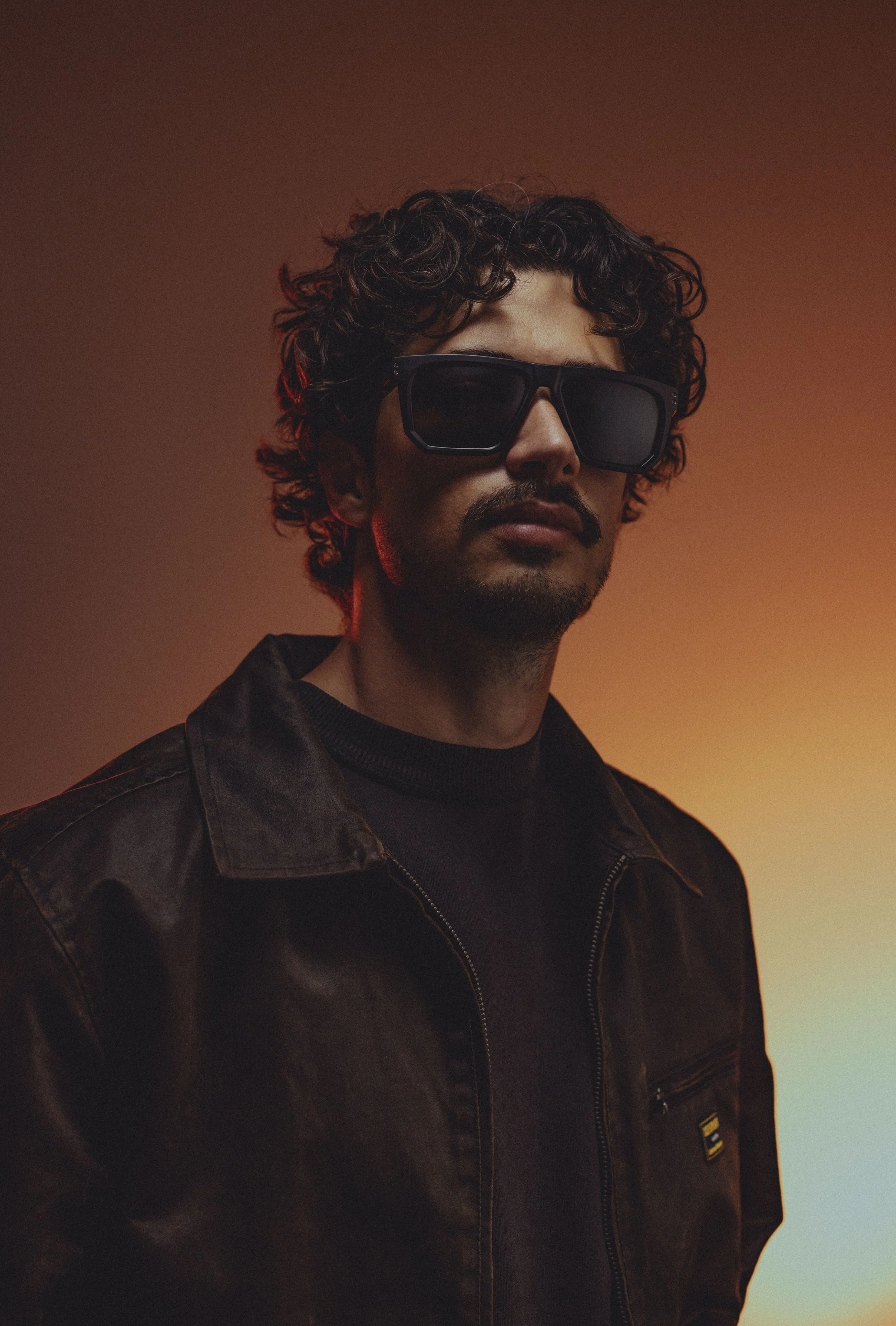 A man with curly hair and sunglasses wearing a leather jacket, standing against a gradient background of warm and cool tones.