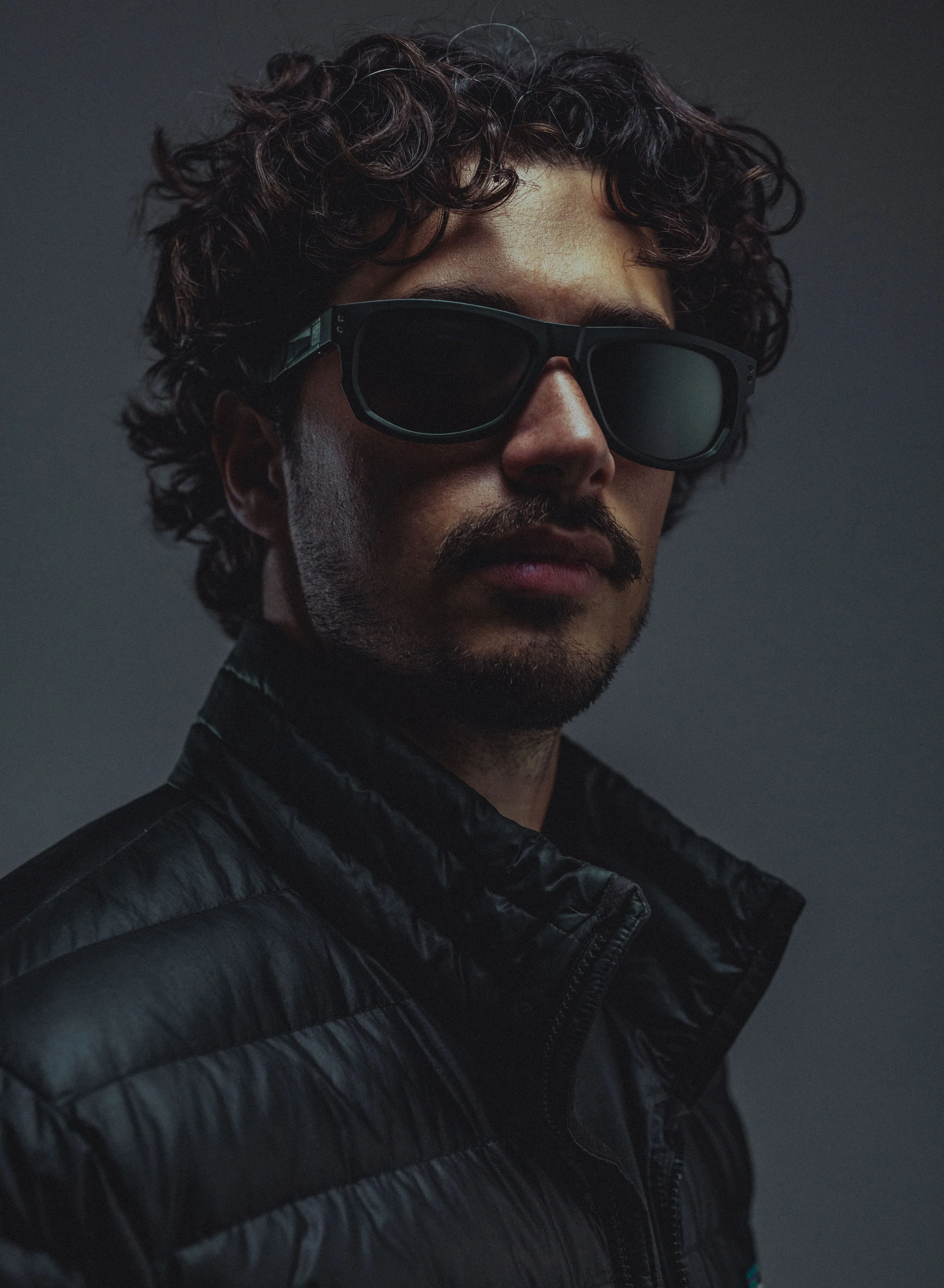 A man with curly dark hair and a beard wearing black sunglasses and a black padded jacket.